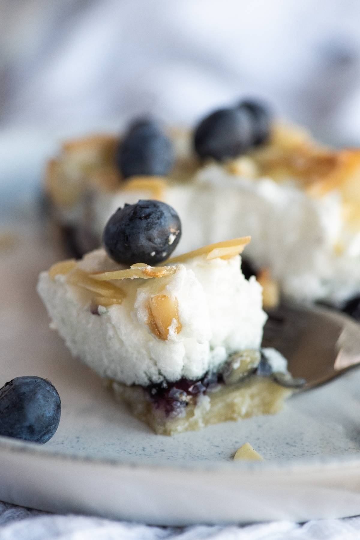 A close-up of a fork holding a bite of cheesecake topped with blueberries and almond slices—an easy blueberry dessert recipe reminiscent of Blueberry Pie Meringue Squares.