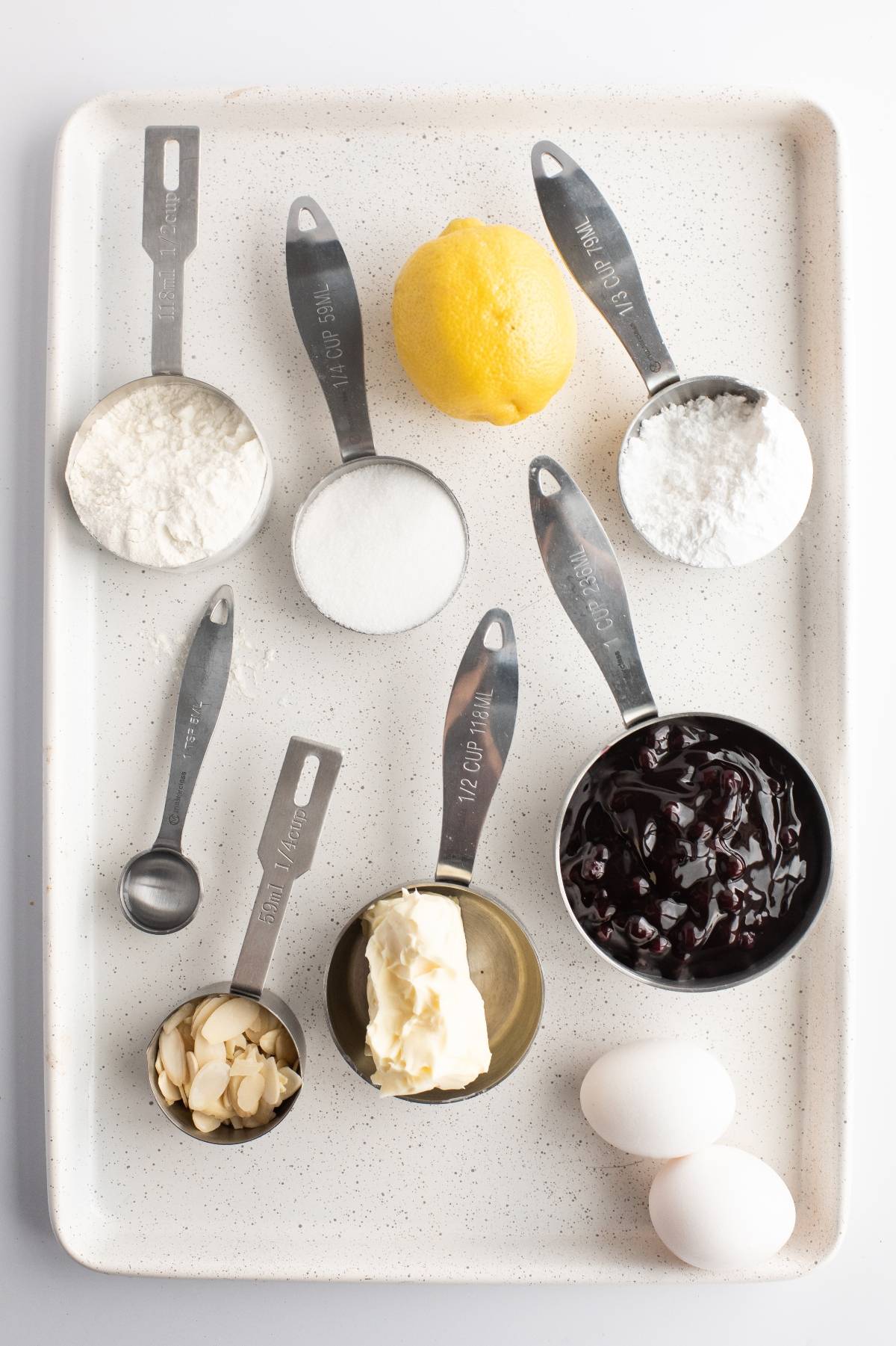 Baking ingredients on a tray: flour, sugar, lemon, butter, jam, eggs, almonds, and powdered sugar in measuring cups—perfect for an almond and blueberry cookie bars recipe.
