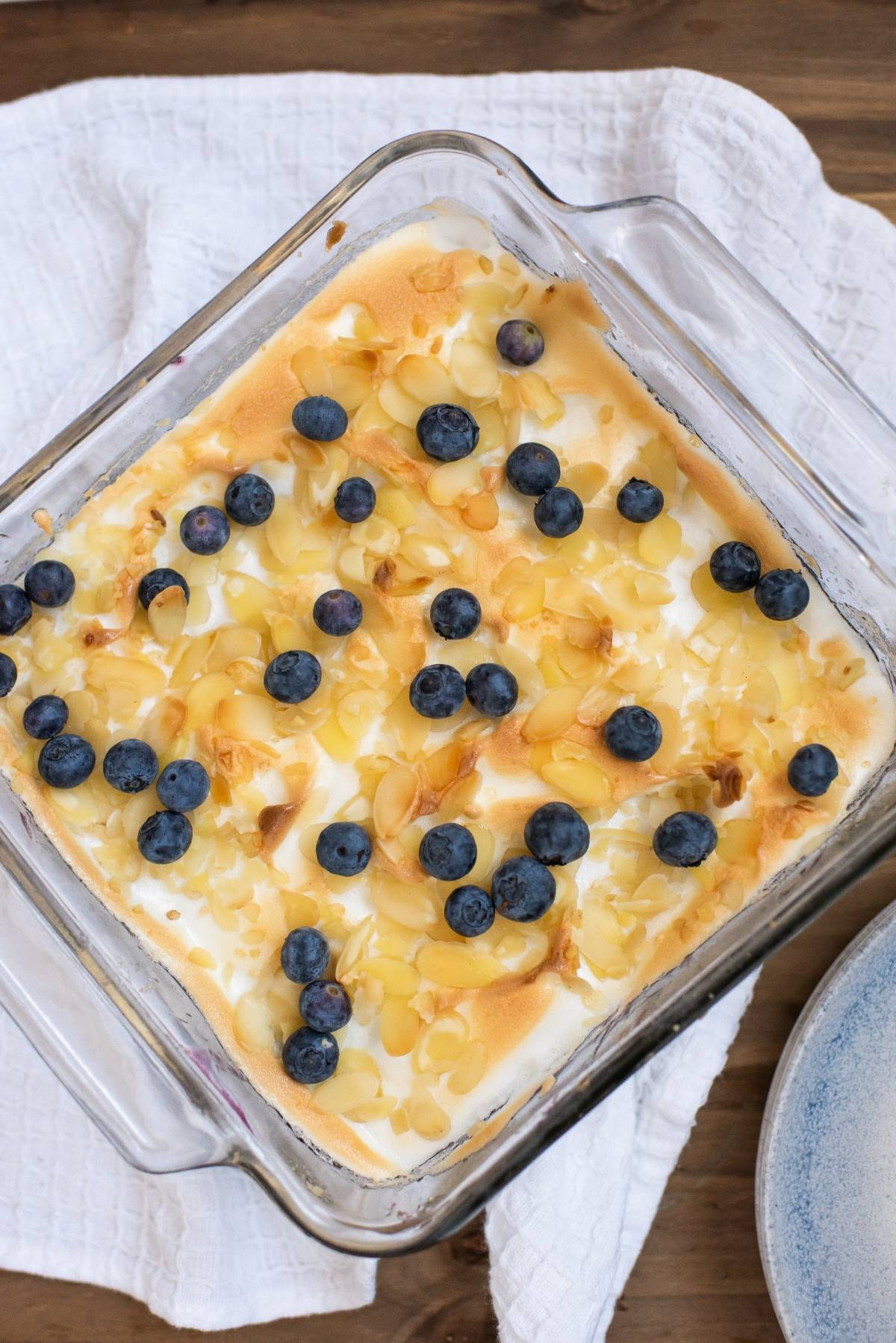 A baked Blueberry Pie Meringue Squares dessert topped with blueberries and almond slices in a glass dish on a white towel—a delicious and easy blueberry dessert recipe for any occasion.
