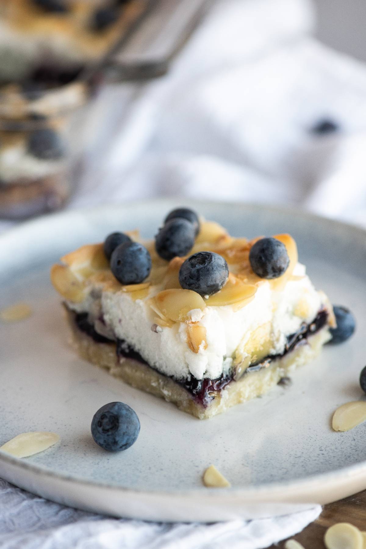 A dessert bar topped with blueberries, almond slices, and a creamy layer, this easy blueberry dessert recipe is served on a plate for a delightful treat.