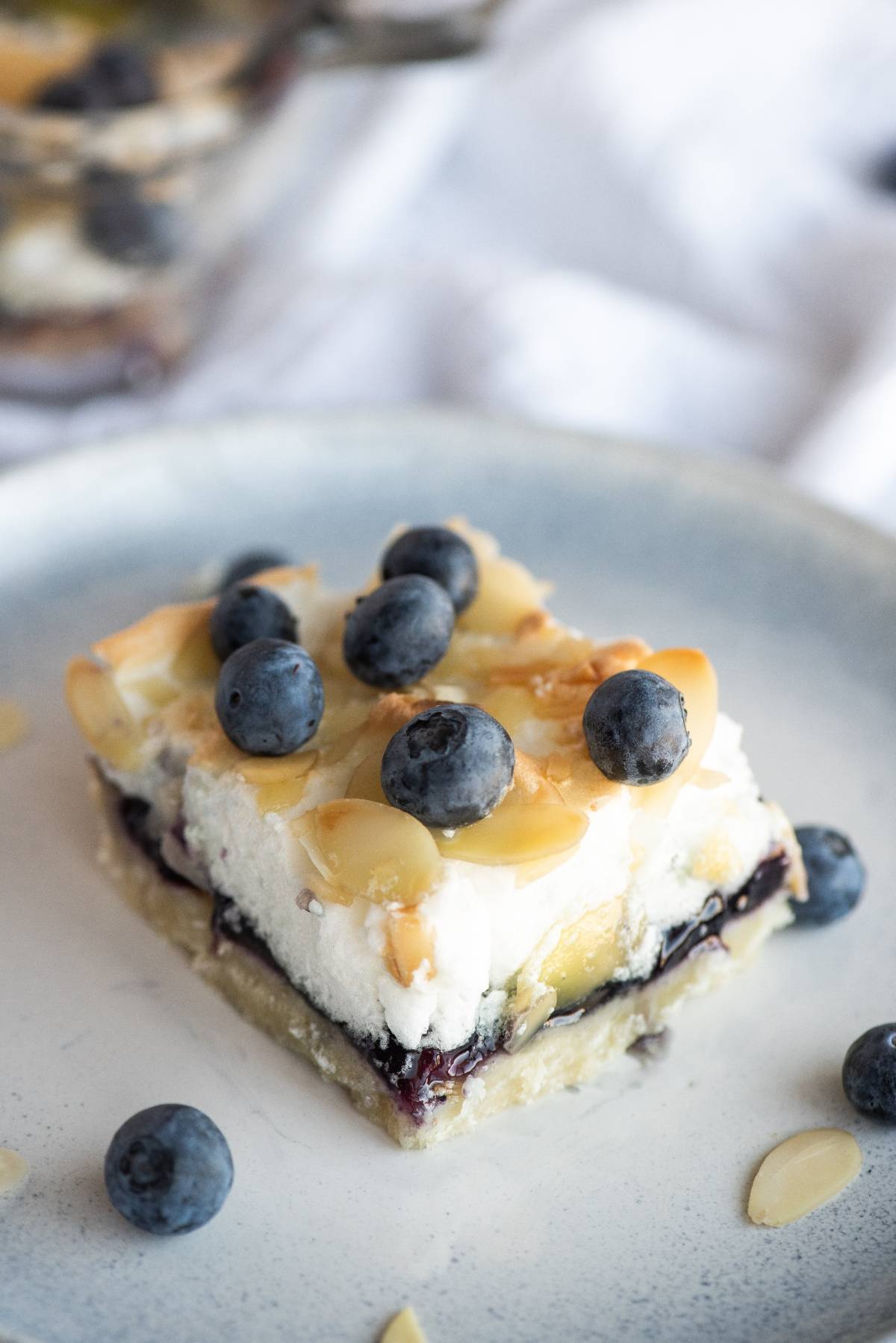 A dessert bar topped with blueberries and almond slices on a plate, with a creamy layer visible—these almond and blueberry cookie bars are a delightful treat.
