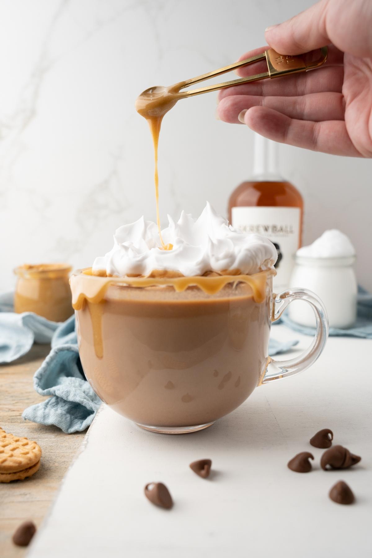 A hand drizzles caramel sauce over a whipped cream topped coffee in a clear mug, creating a tempting treat perfect for any Skrewball whiskey recipe or boozy salted peanut butter hot chocolate.