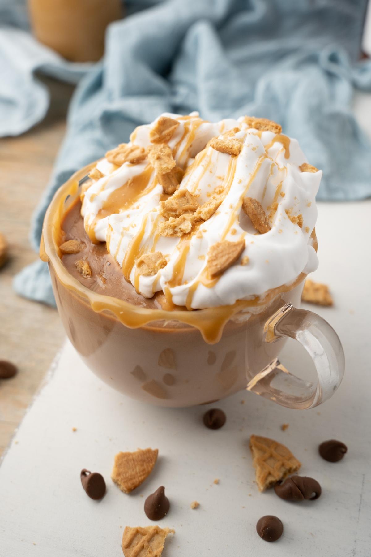 A glass mug of iced coffee topped with whipped cream, caramel drizzle, and cookie crumbles—like a chilled twist on peanut butter hot cocoa.