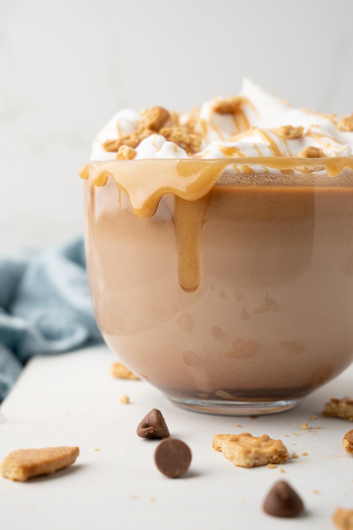 A glass mug of boozy salted peanut butter hot chocolate topped with whipped cream, caramel, and cookie crumbs sits on a white surface.