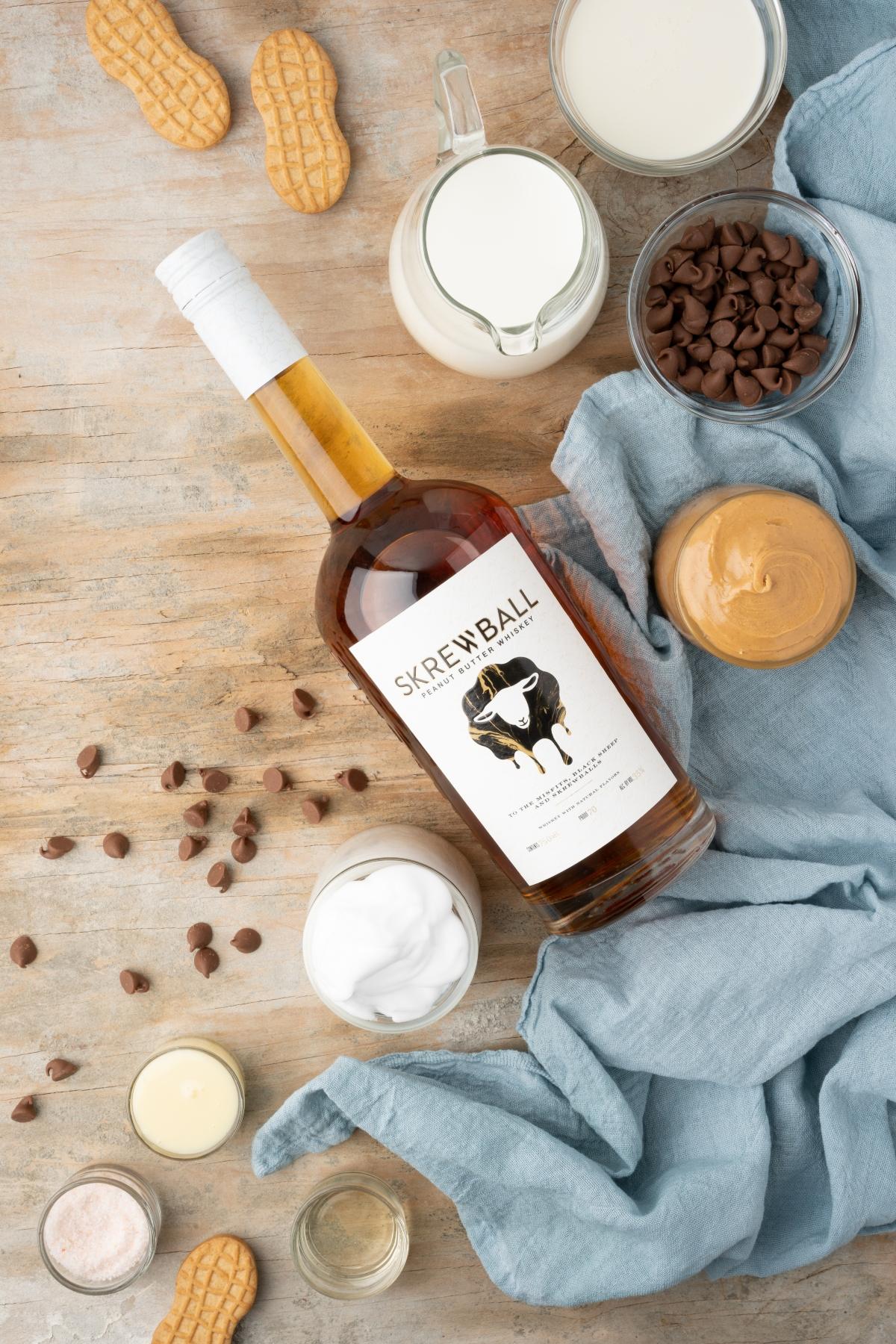 Bottle of Skrewball whiskey surrounded by peanut butter, chocolate chips, milk, and peanuts on a wooden surface—perfect for crafting a rich peanut butter hot cocoa or your next cozy Skrewball whiskey recipe.