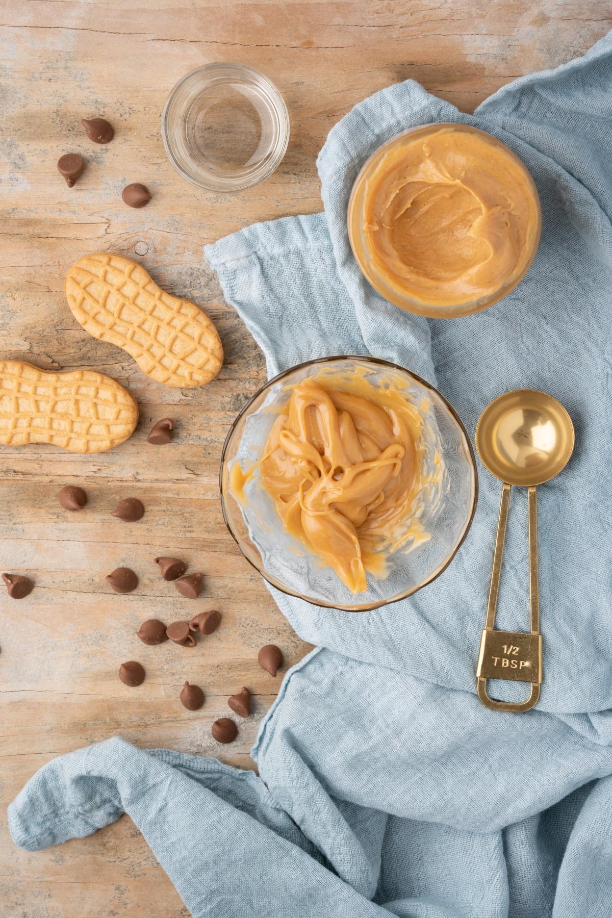 Peanut butter in bowls, cookies, chocolate chips, and a measuring spoon on a wooden surface with a blue cloth—perfect for whipping up a Boozy Salted Peanut Butter Hot Chocolate or a rich Skrewball whiskey recipe.