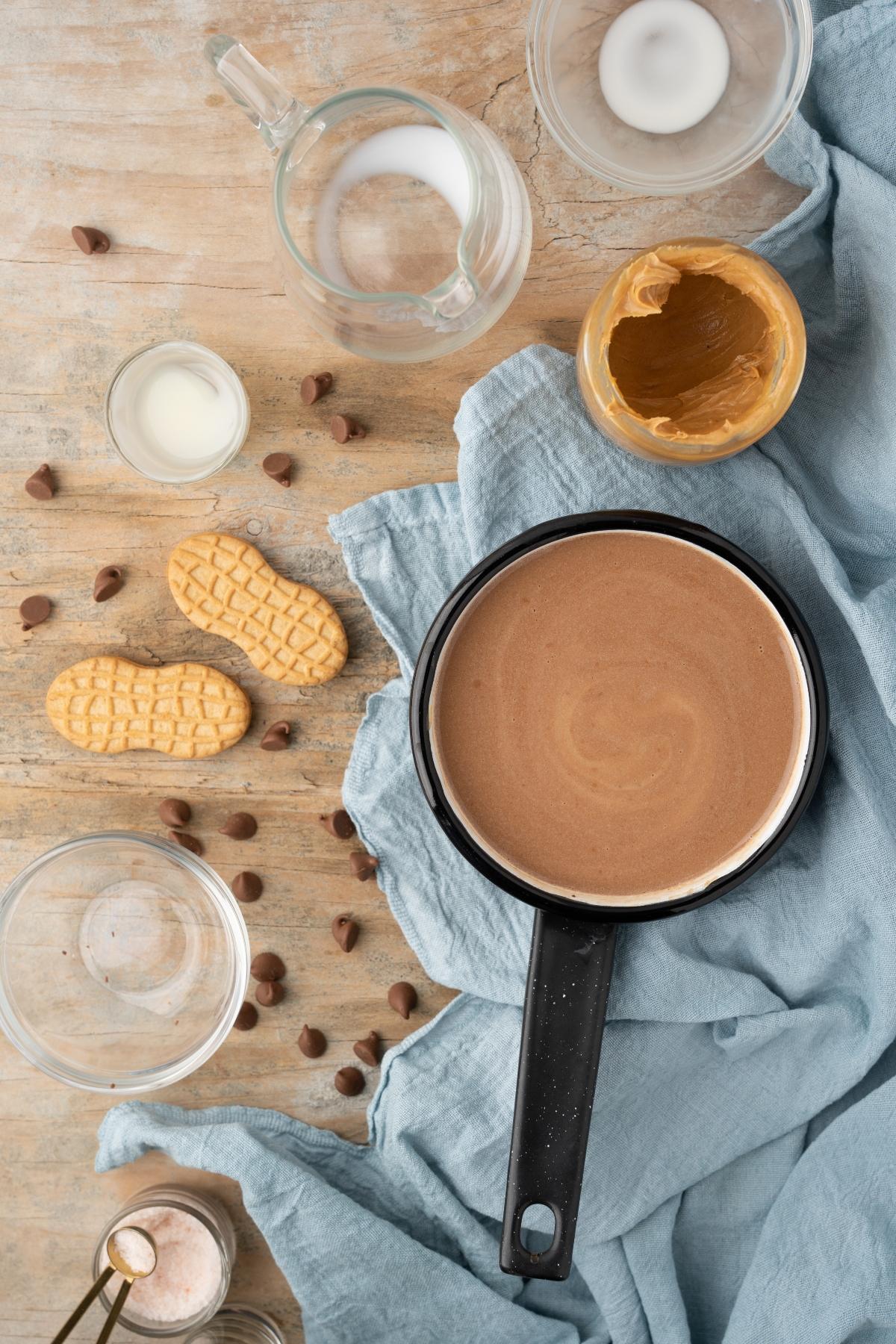 A saucepan of chocolate mixture with cookies, peanut butter, chocolate chips, and milk on a wooden table—perfect for making decadent Boozy Salted Peanut Butter Hot Chocolate.