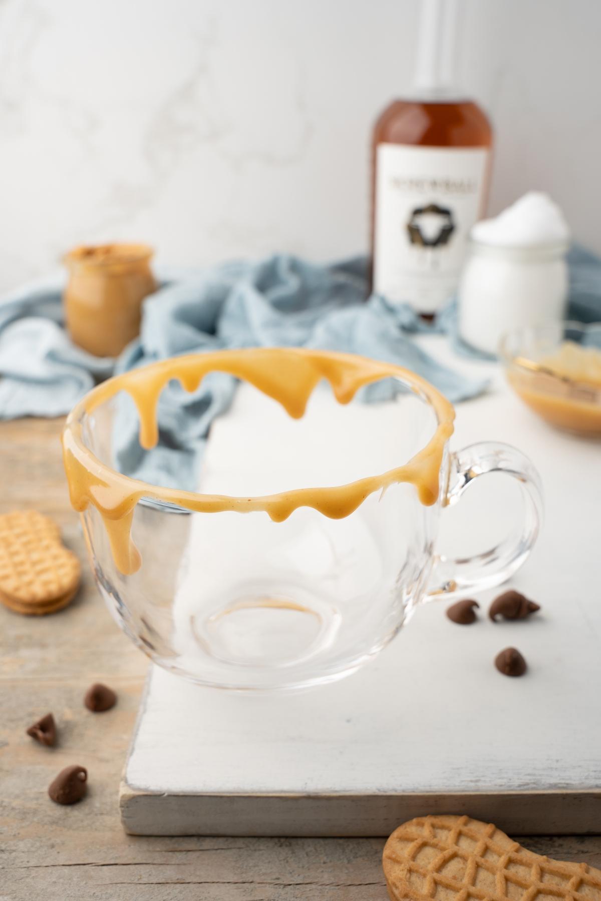 Clear glass mug with peanut butter dripped on the rim, surrounded by cookies and dessert ingredients—a tempting setting for hot chocolate and whiskey or a cozy peanut butter hot cocoa.