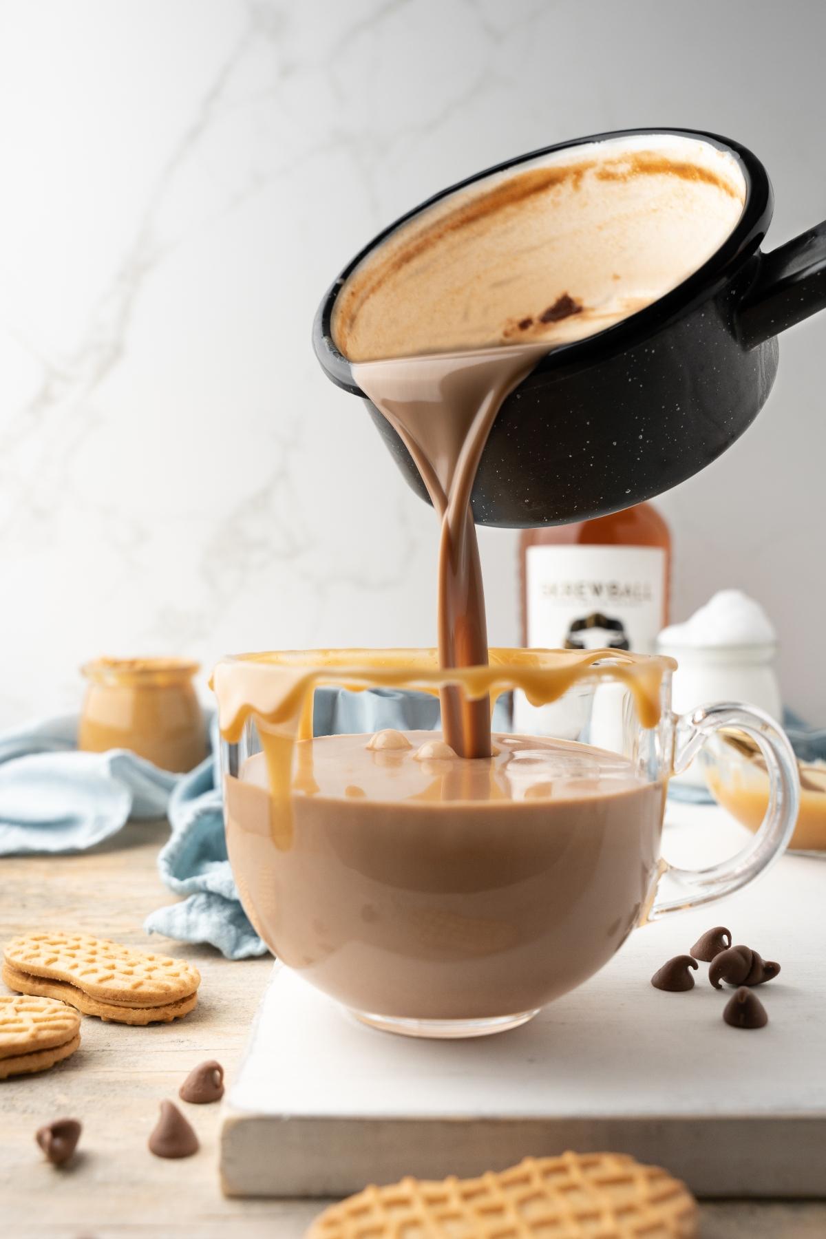 A pot pours creamy peanut butter hot cocoa into a glass mug, with cookies and chocolate chips nearby.
