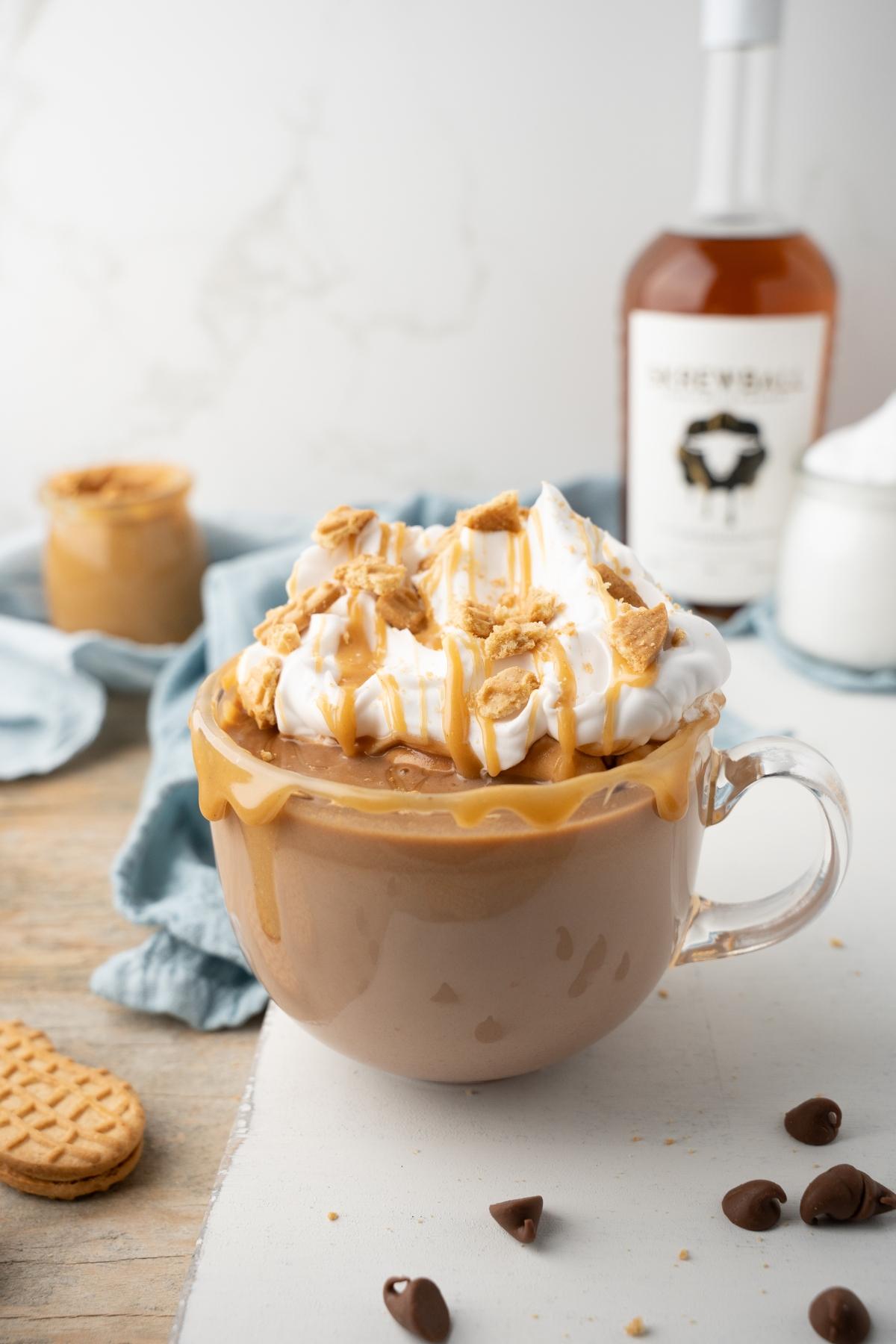 A glass mug of hot peanut butter drink topped with whipped cream, caramel drizzle, and cookie crumbles.