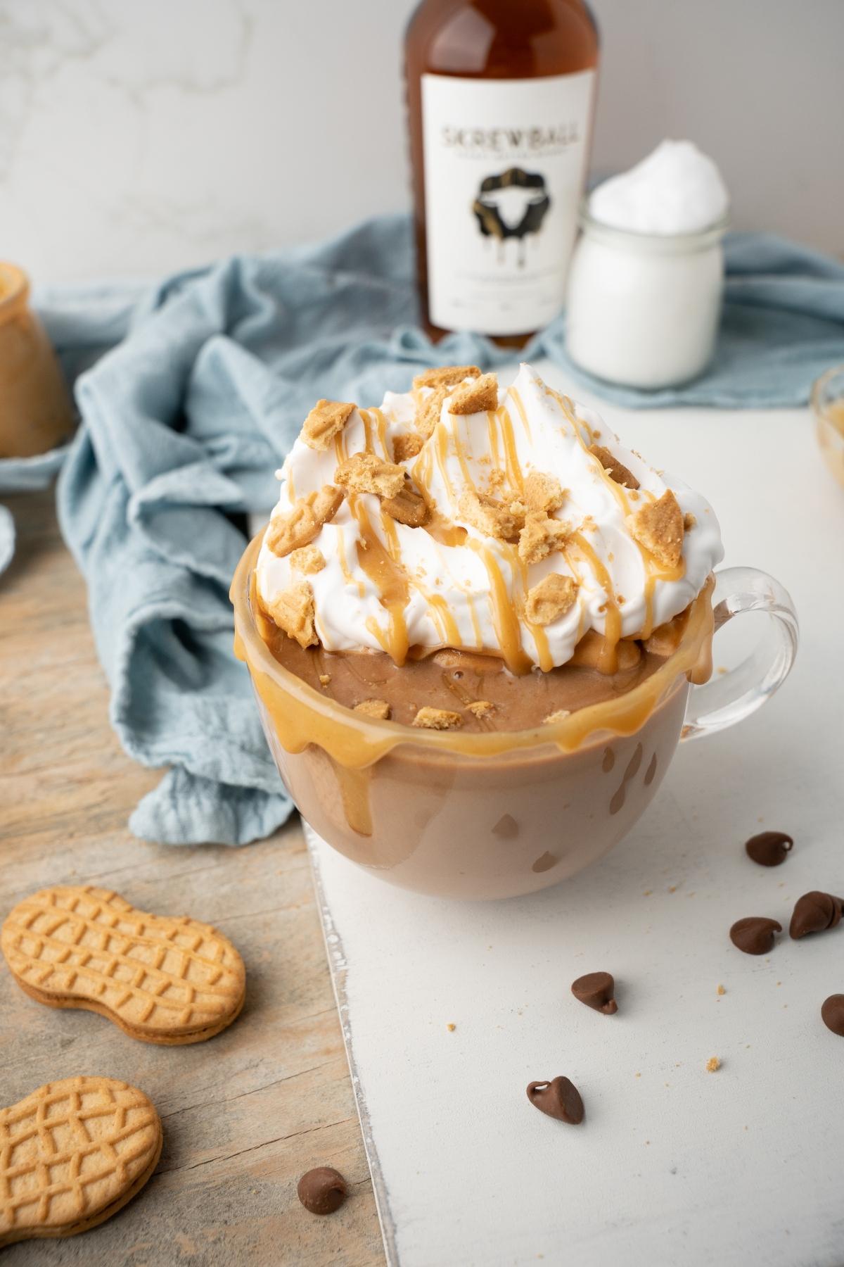 A mug of creamy Boozy Salted Peanut Butter Hot Chocolate, topped with whipped cream, caramel, and cookie crumbs, sits on a table.