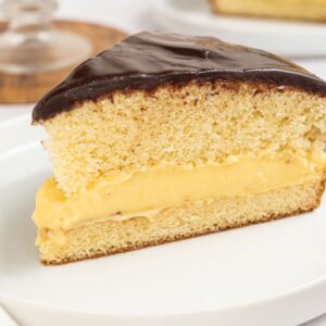 A delicious slice of Boston cream pie with rich chocolate glaze and creamy custard filling sits elegantly on a white plate.