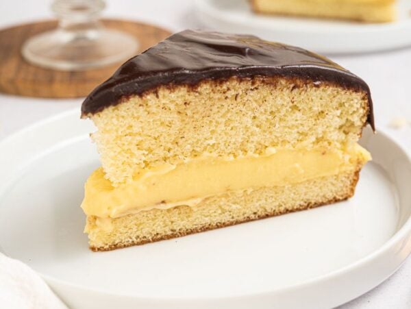 A delicious slice of Boston cream pie with rich chocolate glaze and creamy custard filling sits elegantly on a white plate.