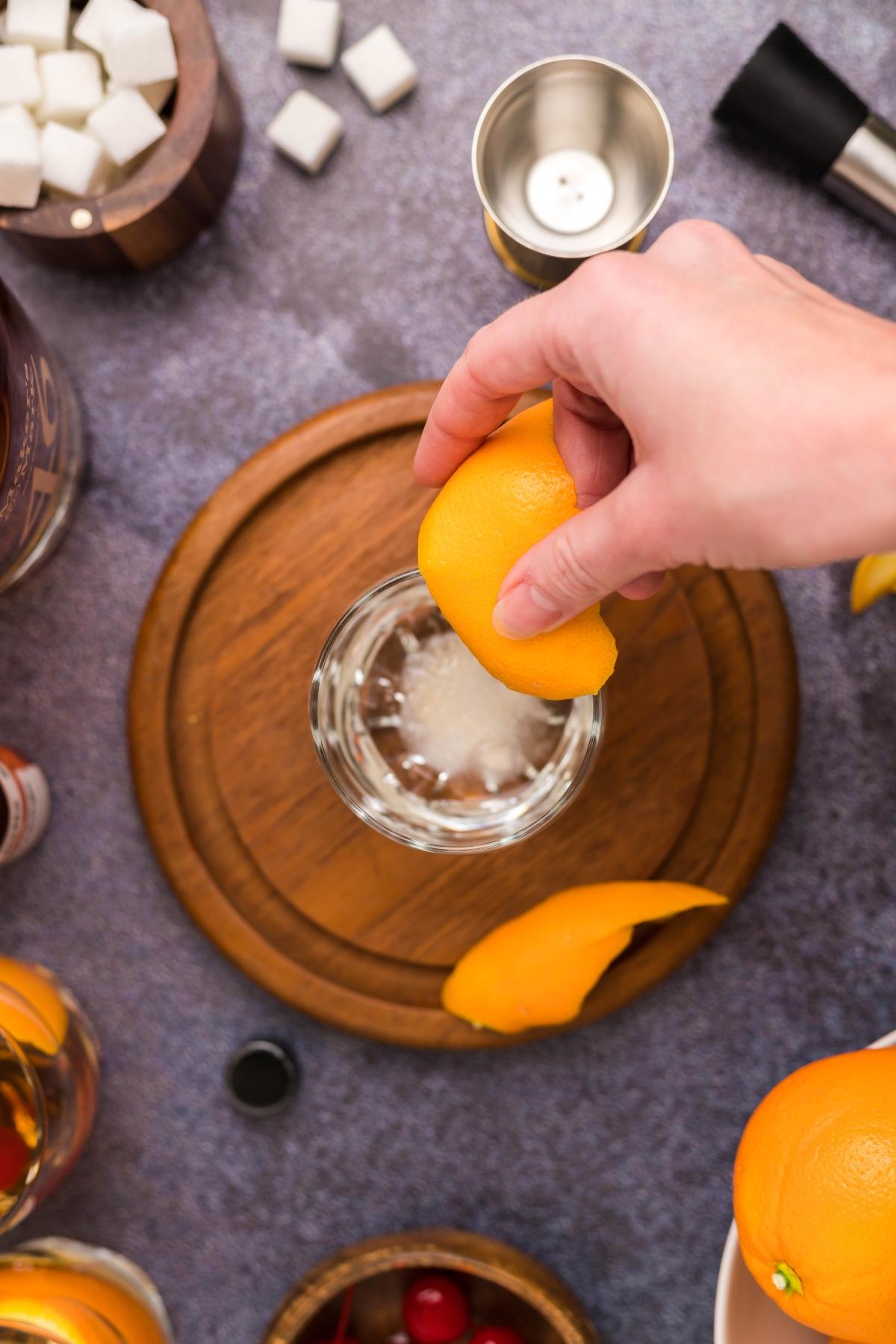 A hand places an orange peel into a glass on a wooden board, surrounded by cocktail ingredients—a classic bourbon cocktail in the making.