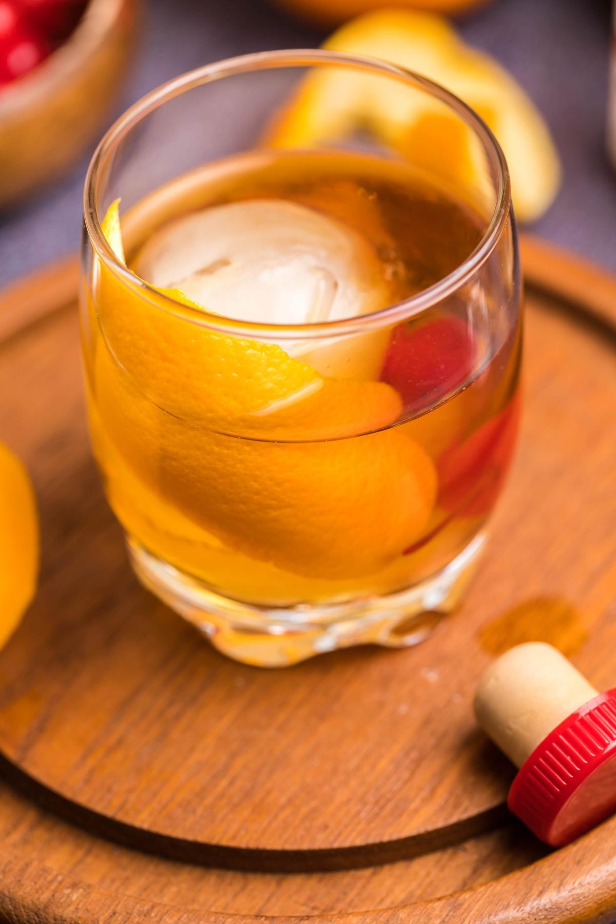 A glass of cocktail with ice, orange peel, and cherries on a wooden board hints at the best old fashioned recipe—a timeless drink you can make with bourbon. A bottle cap rests nearby, completing the classic scene.