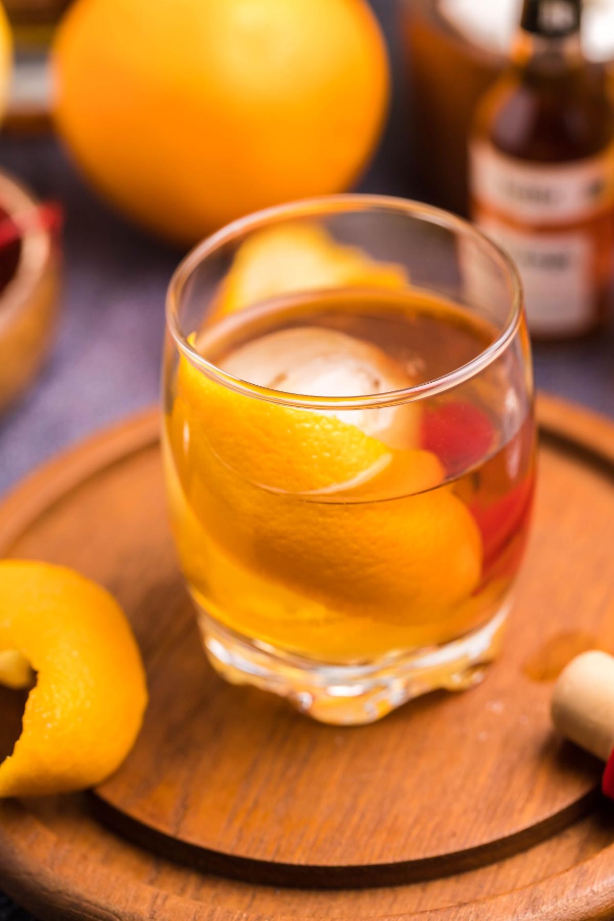 A glass of classic bourbon cocktail with an orange peel and ice on a wooden tray, surrounded by citrus and bitters bottles—perfect for those wanting to learn how to make a Bourbon old fashioned.