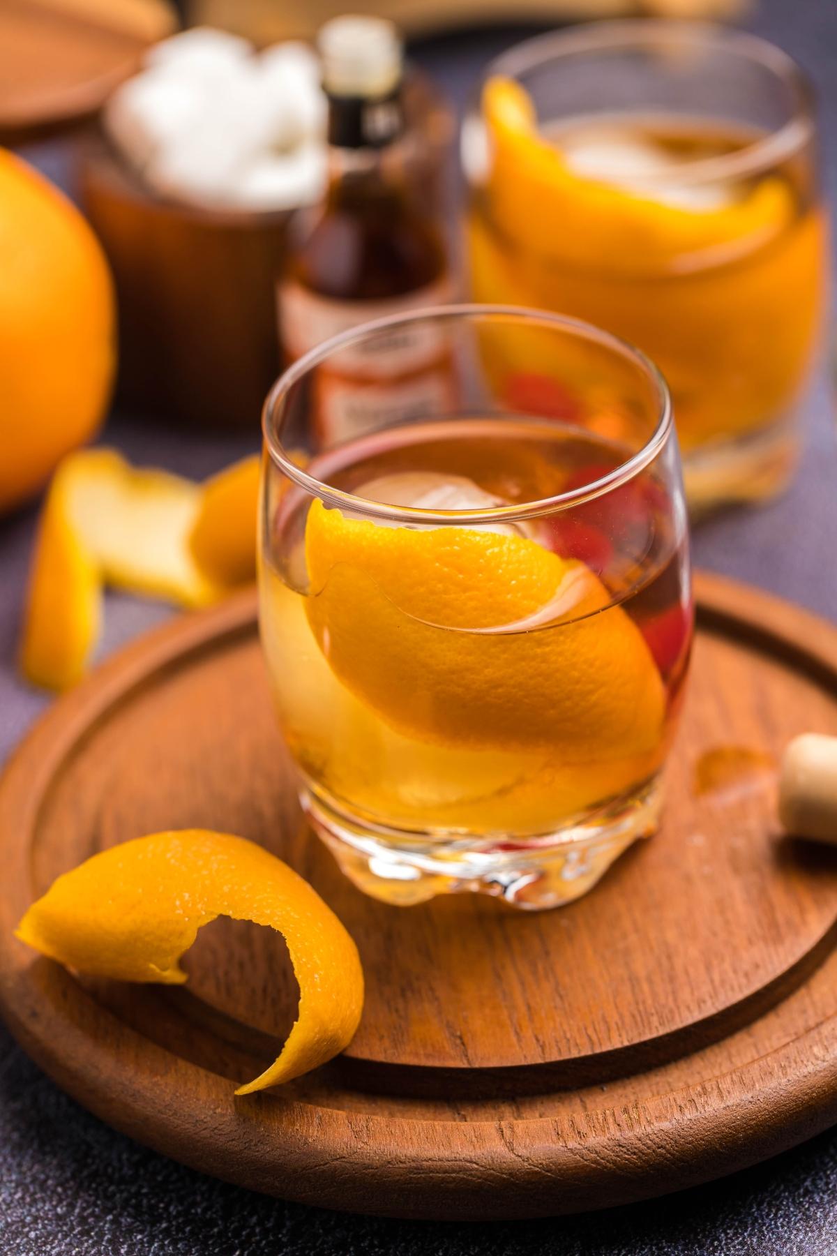 A glass of whiskey with an orange peel garnish on a wooden tray, showcasing a classic bourbon cocktail, with another drink and ingredients in the background.
