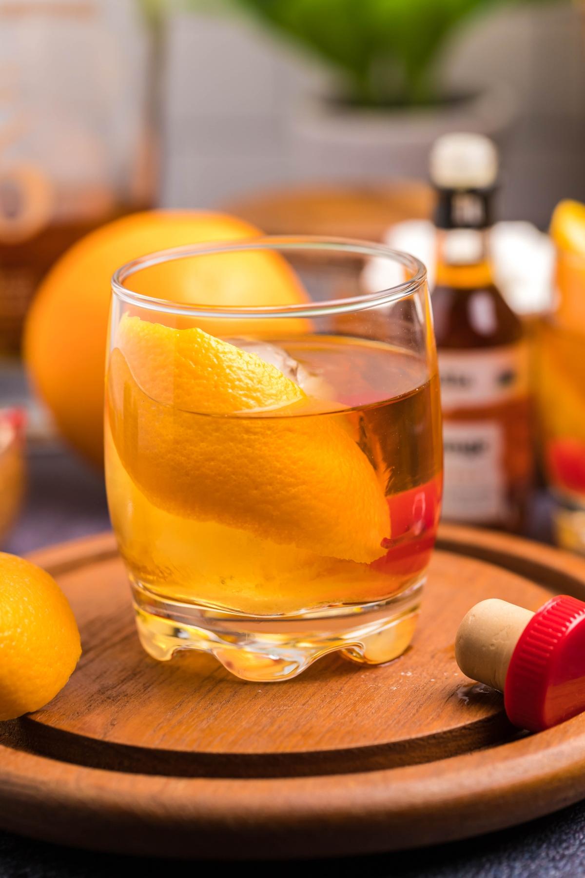 A glass of Bourbon Old Fashioned with an orange peel and cherry sits on a wooden tray, surrounded by lemons and bitters bottles—the perfect drink you can make with bourbon.