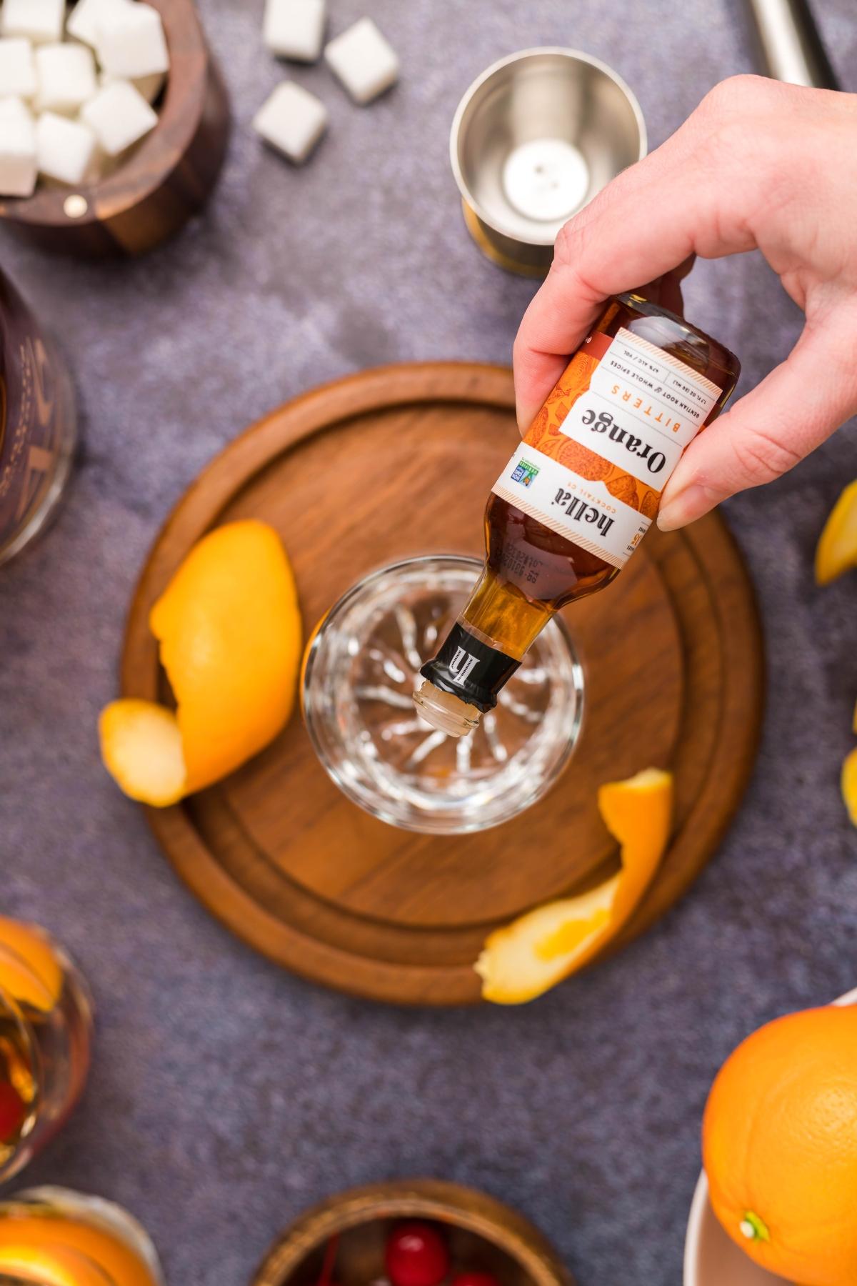 A hand pours orange bitters into a glass on a wooden tray, surrounded by orange peels and sugar cubes—the essential steps for how to make a bourbon old fashioned, the classic bourbon cocktail.