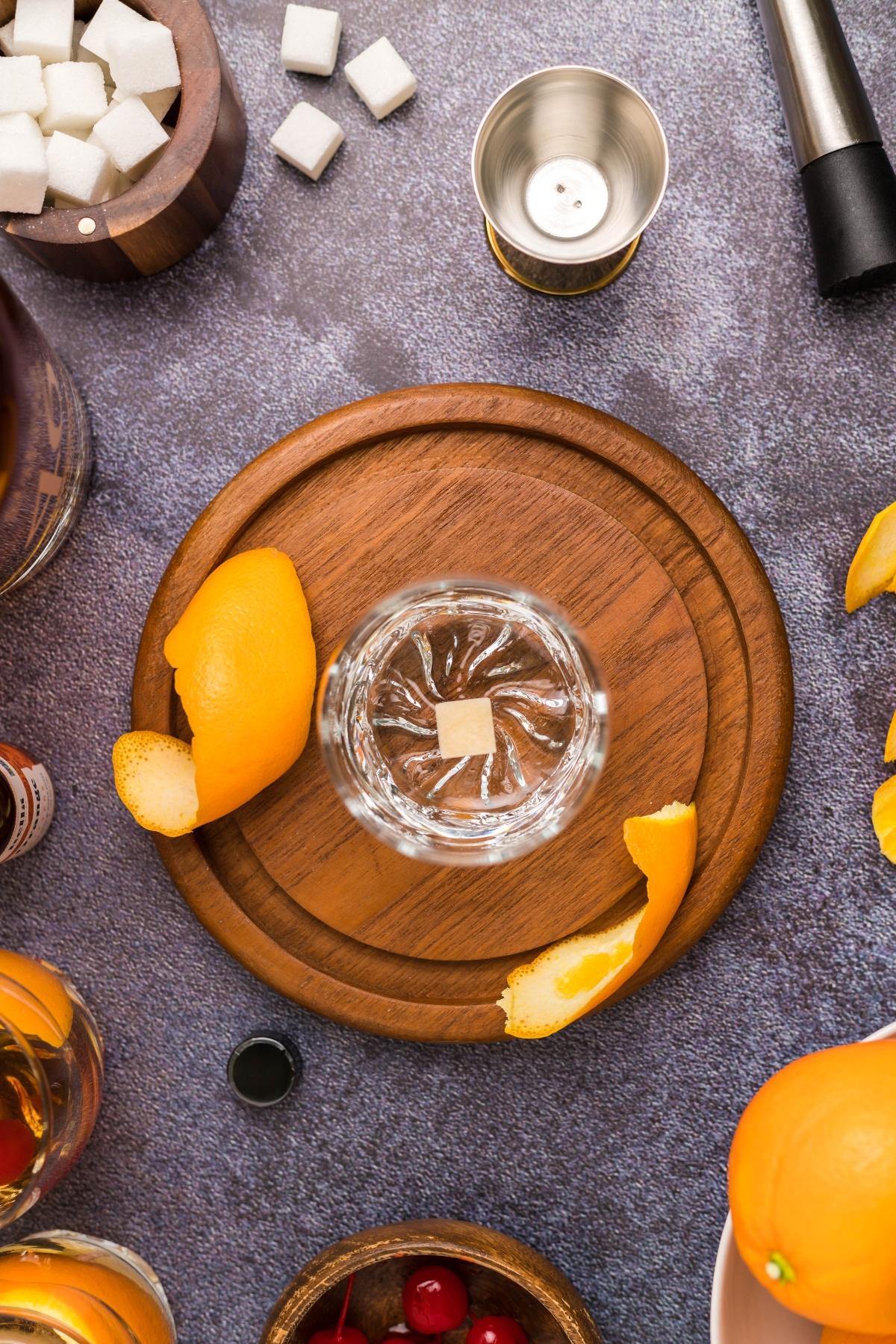 Overhead view of a glass with a sugar cube and orange peel on a wooden board, surrounded by ingredients for the best old fashioned recipe—a classic drink you can make with bourbon.