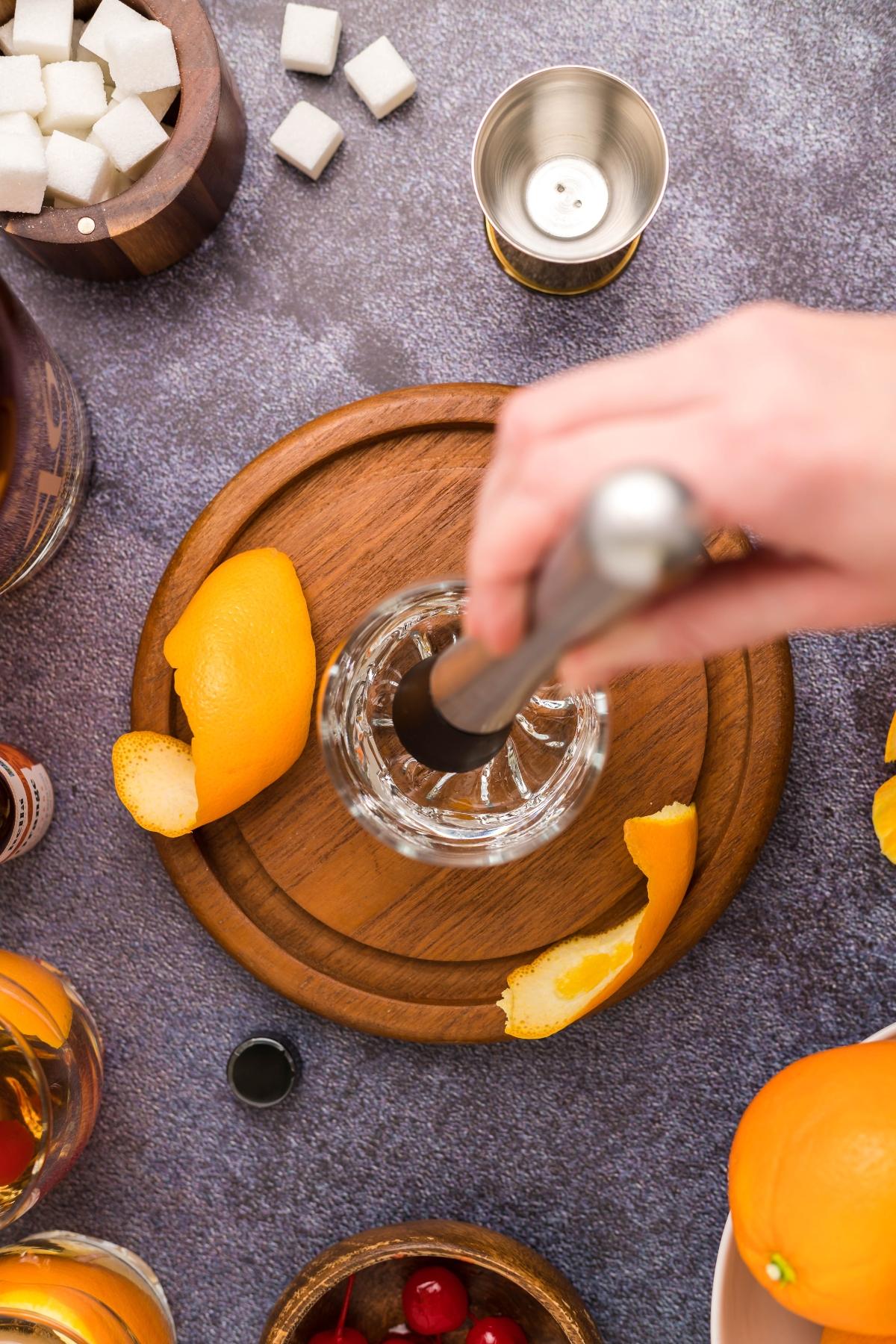 A hand muddling ingredients in a glass with orange peel, surrounded by cocktail-making items—capturing the timeless process of creating a classic bourbon cocktail.