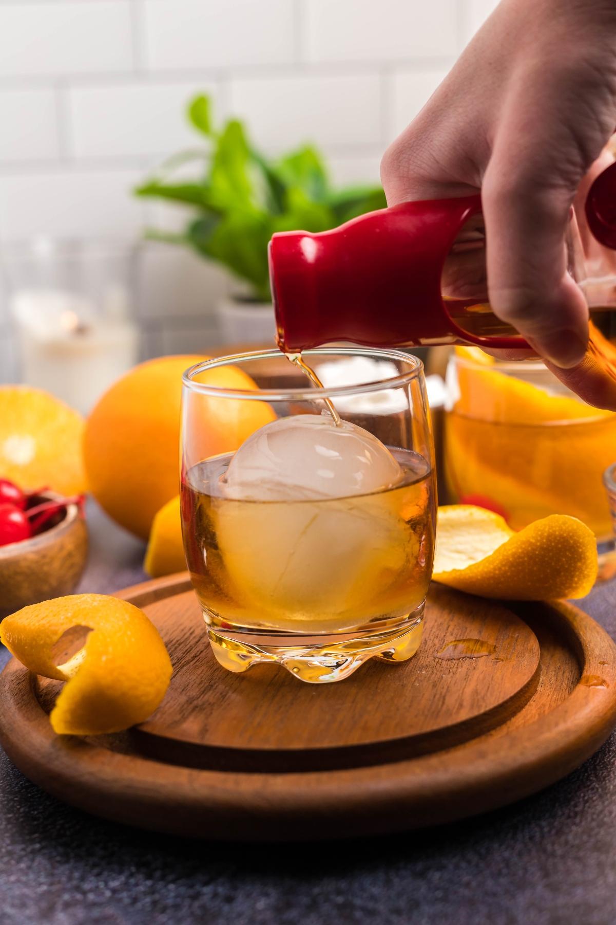 A hand pours whiskey into a glass with a round ice cube, surrounded by citrus and cherries—the perfect way to enjoy a classic bourbon cocktail.