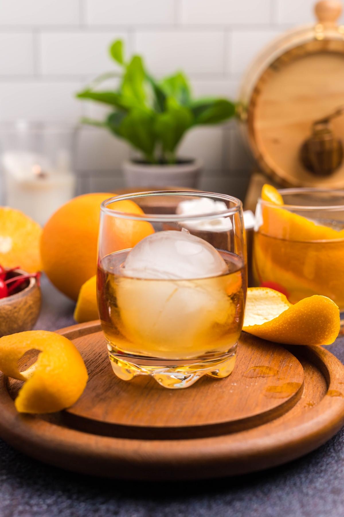 Glass of whiskey with a round ice cube, orange peel garnish, and oranges on a wooden tray—perfect for those seeking the best old fashioned recipe.