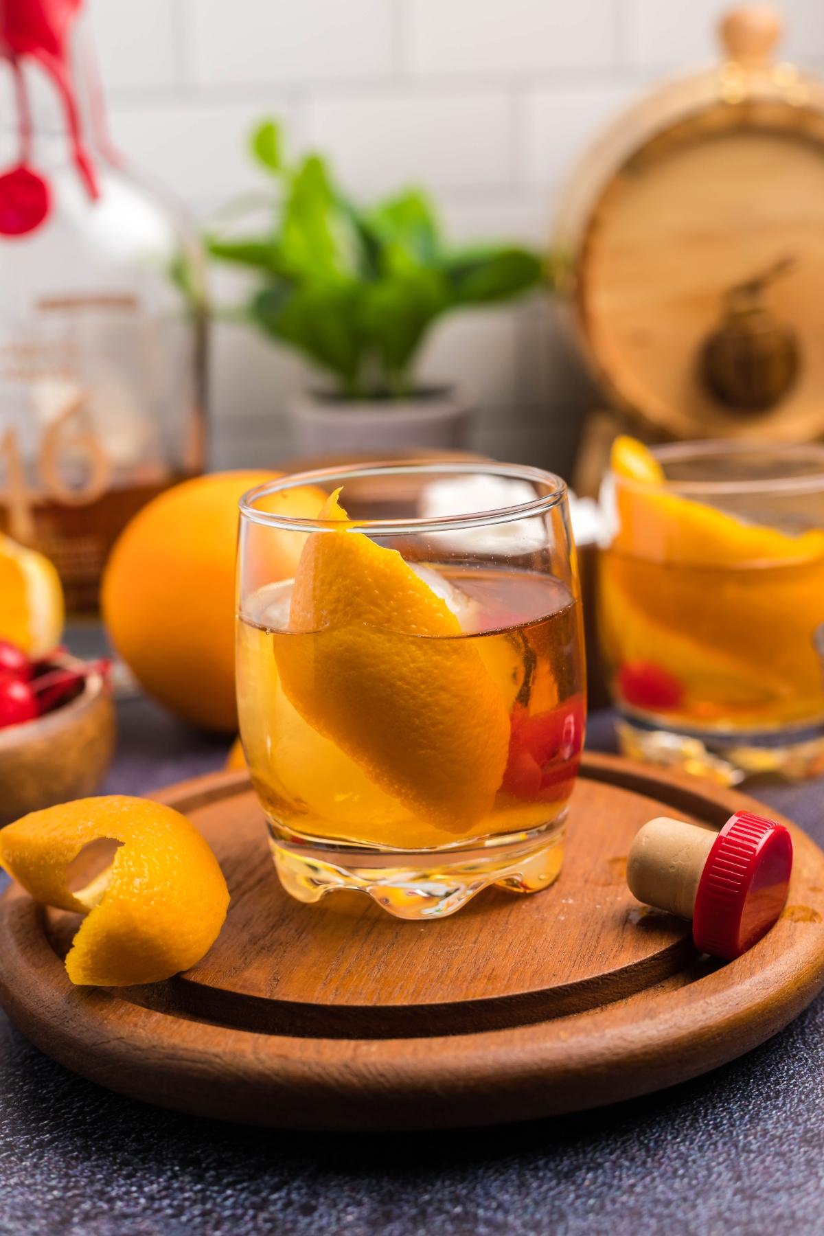 Glass of whiskey with orange peel and cherry garnish on a wooden board, showing how to make a bourbon old fashioned—one classic drink you can make with bourbon. Fruit and bottle in the background enhance the timeless appeal.