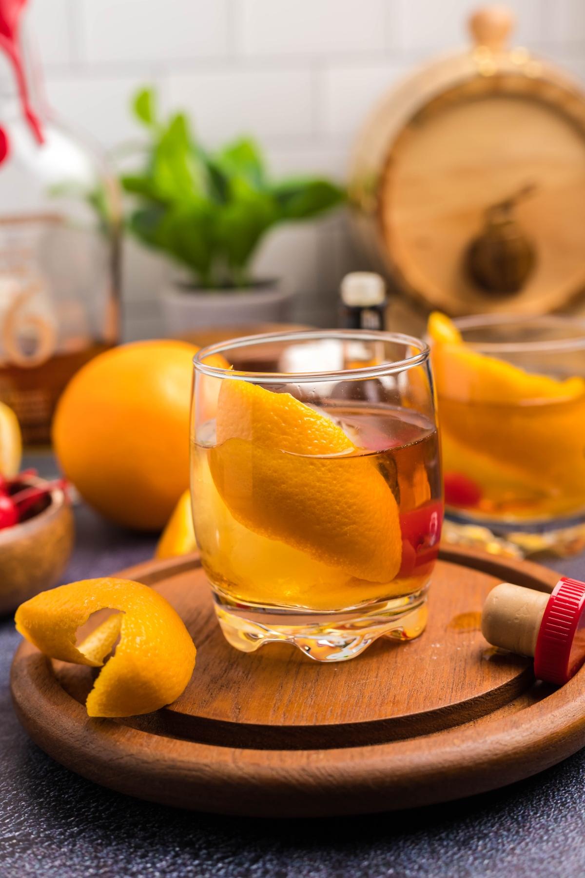 A glass of whiskey with an orange peel garnish sits on a wooden tray, surrounded by fruit and bar items—perfect for those wanting to enjoy a classic bourbon cocktail or learn how to make a bourbon old fashioned at home.