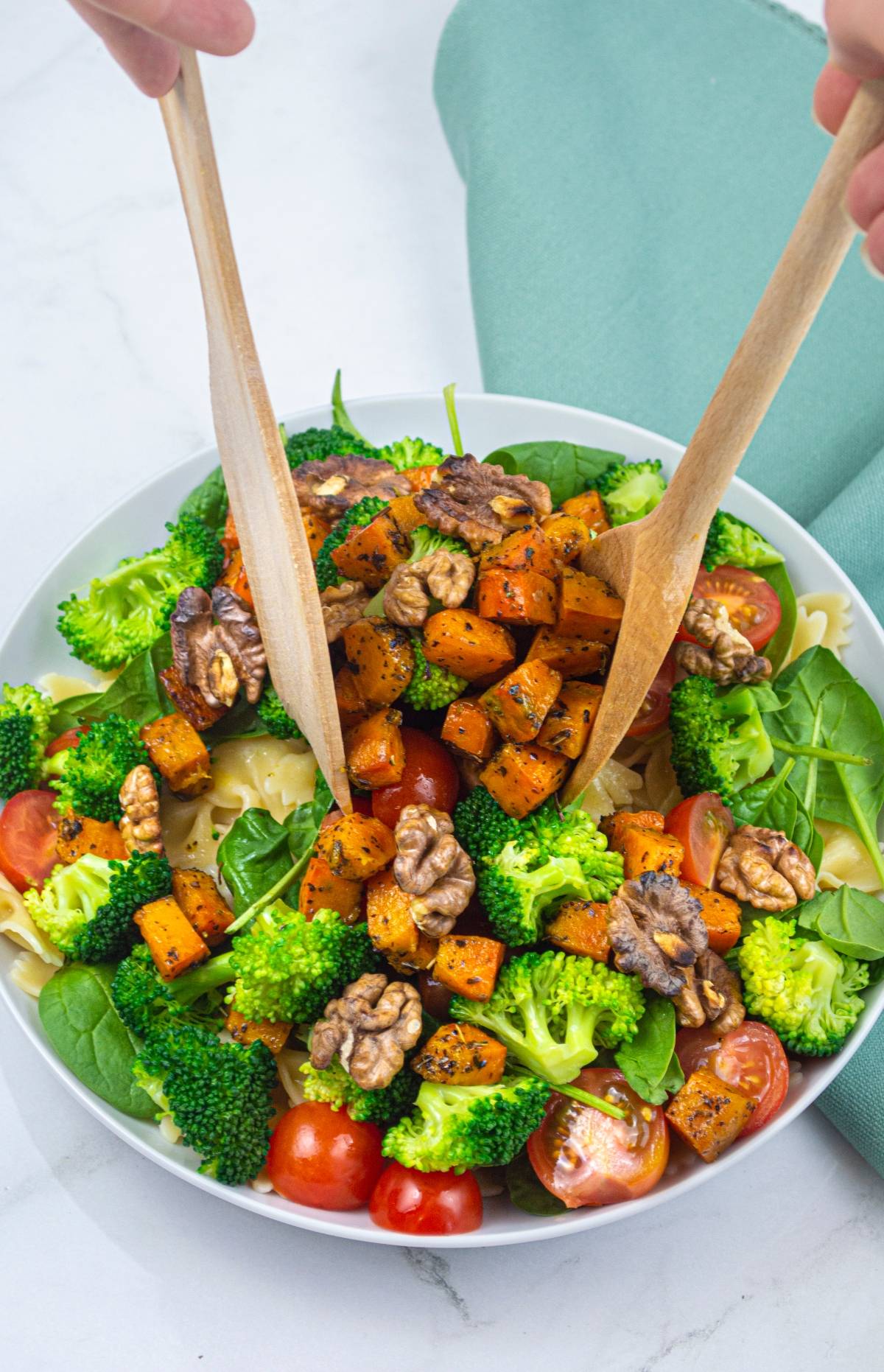 A vibrant roasted squash salad with spinach, broccoli, cherry tomatoes, and walnuts is tossed with wooden utensils—a fresh twist on a classic Butternut Squash and Broccoli Pasta Salad.