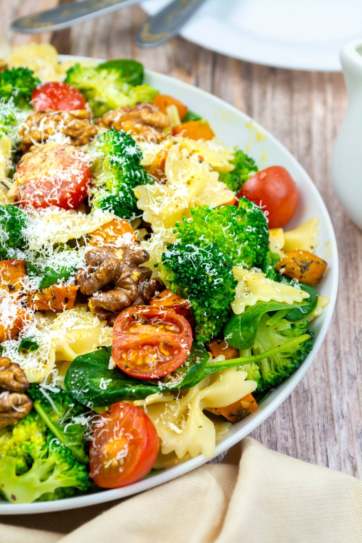 Colorful Vegetable pasta salad with broccoli, cherry tomatoes, spinach, walnuts, and grated cheese in a white bowl.