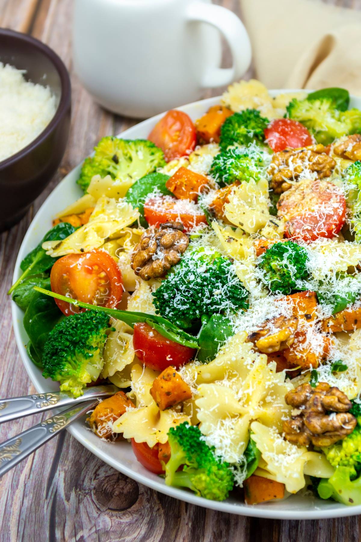 A plate of vibrant vegetable pasta salad with bowtie pasta, broccoli, cherry tomatoes, walnuts, spinach, and grated cheese on top.