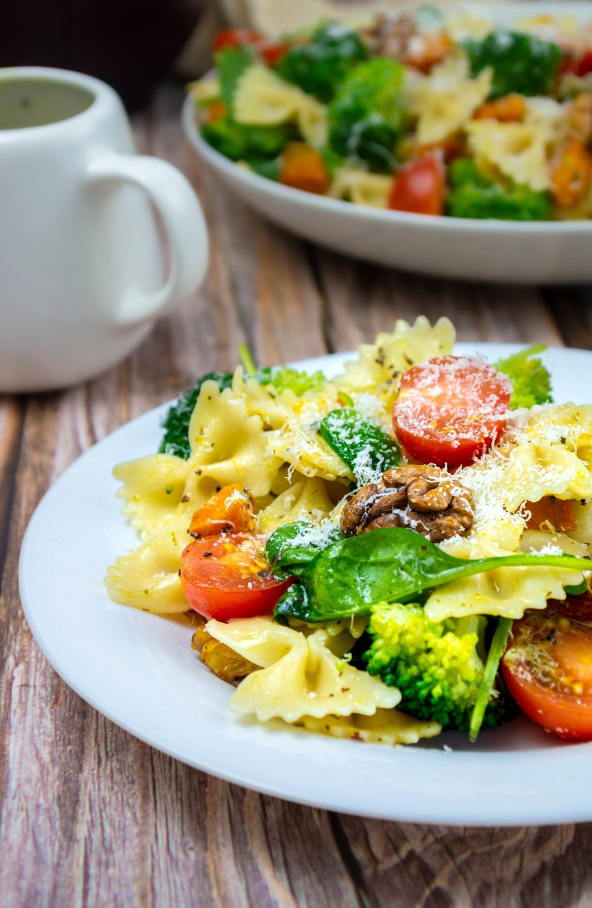 A plate of Vegetable pasta salad with bow tie pasta, broccoli, cherry tomatoes, walnuts, and spinach sits on a wooden table.