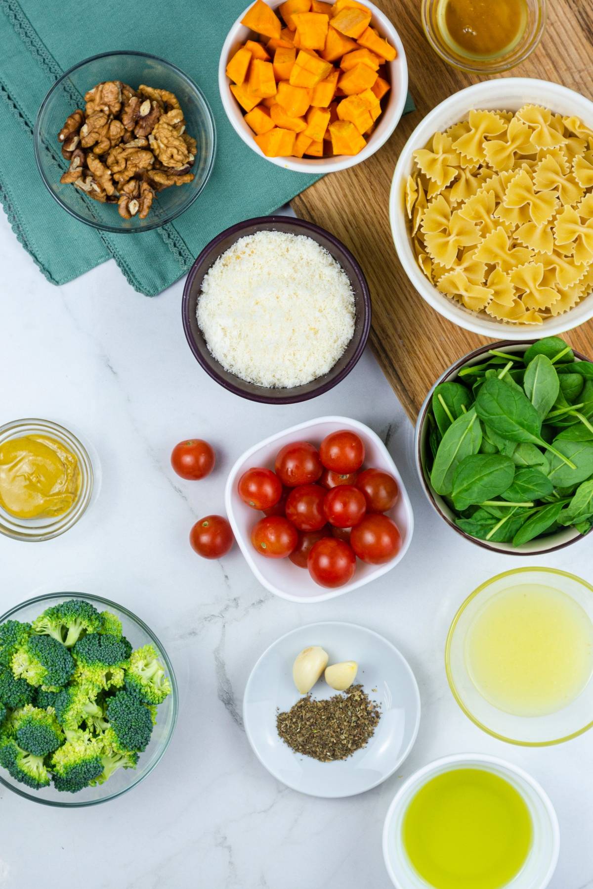 Bowls of pasta, spinach, cherry tomatoes, sweet potatoes, broccoli, walnuts, cheese, oil, garlic, and seasoning create a vibrant Butternut Squash and Broccoli Pasta Salad bursting with fresh flavor.