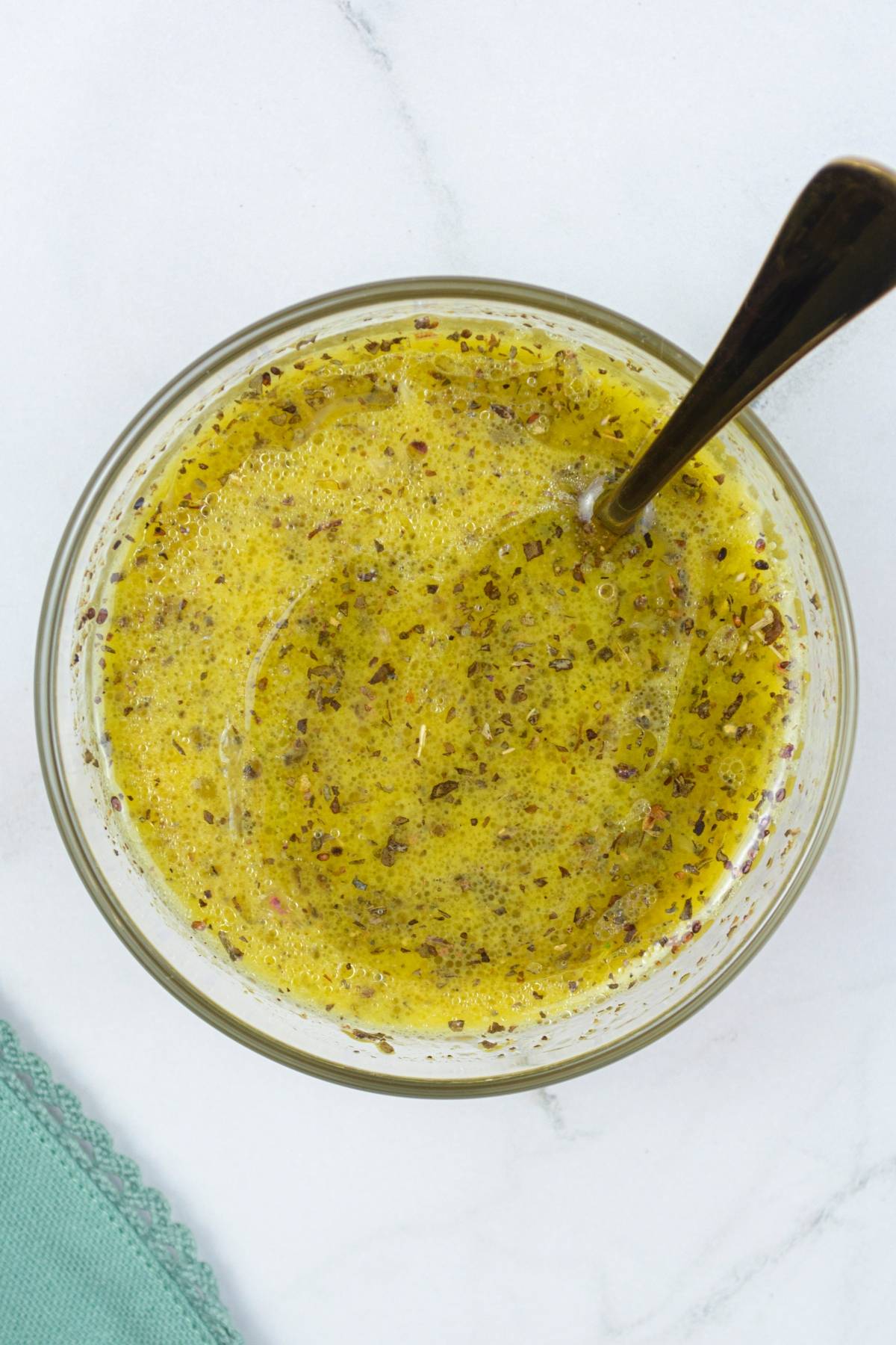 A bowl of Italian salad dressing with herbs and a spoon, perfect for drizzling over a Roasted Squash Salad or a Gluten Free Vegan Pasta on a white surface.