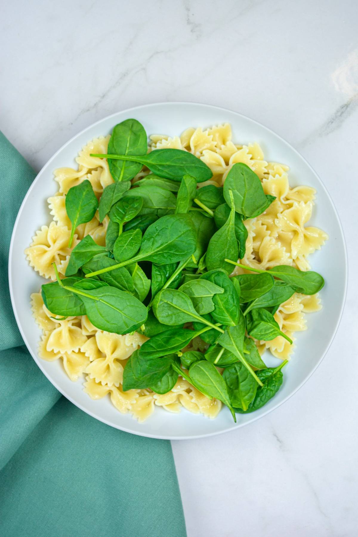 White plate with Gluten free vegan pasta topped with fresh spinach leaves on a light surface, next to a green napkin.