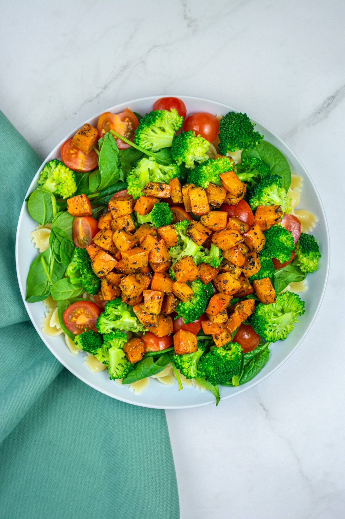 A vibrant vegetable pasta salad featuring spinach, broccoli, cherry tomatoes, gluten free vegan pasta, and roasted sweet potatoes on a white plate.