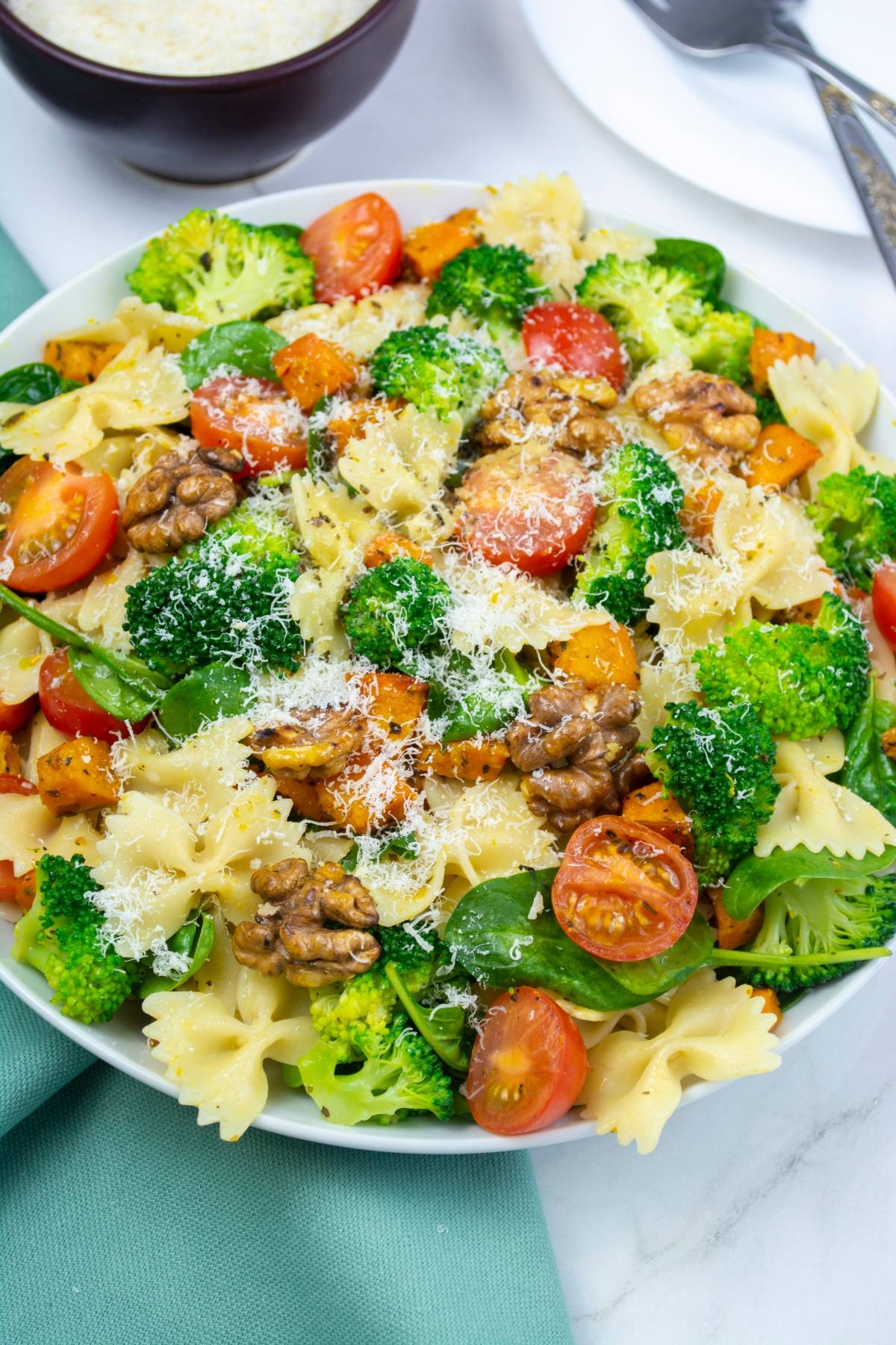 A colorful vegetable pasta salad featuring bow-tie pasta, broccoli, cherry tomatoes, walnuts, and grated cheese served on a plate.