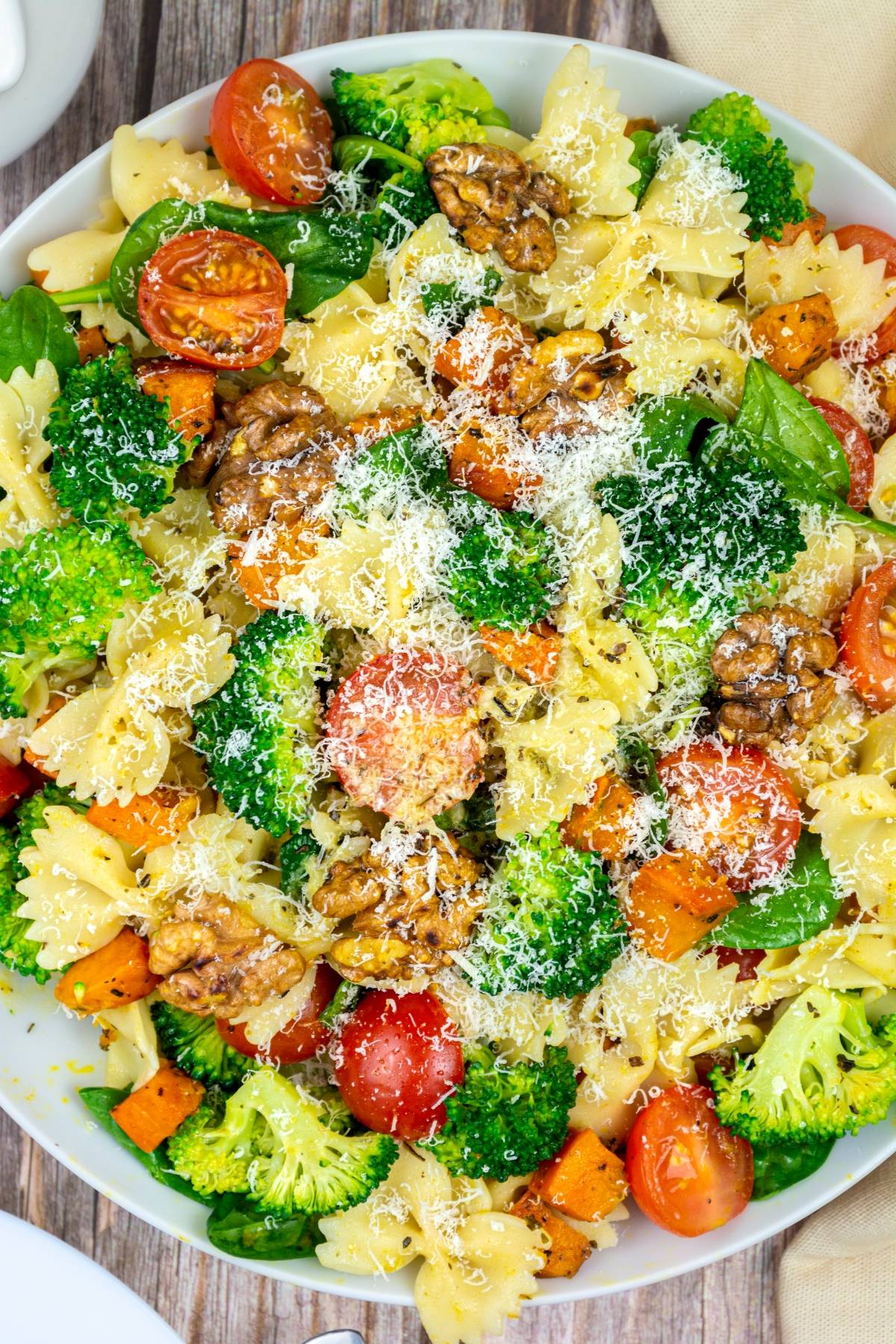 A bowl of vegetable pasta salad with broccoli, cherry tomatoes, walnuts, spinach, and grated cheese on top.