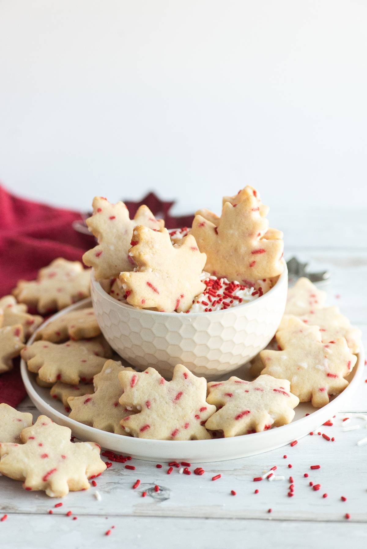 Maple leaf-shaped cookies with red sprinkles arranged in a bowl and on a white plate—perfect cookies to celebrate Canada Day or inspire recipes for a Canada Day party.