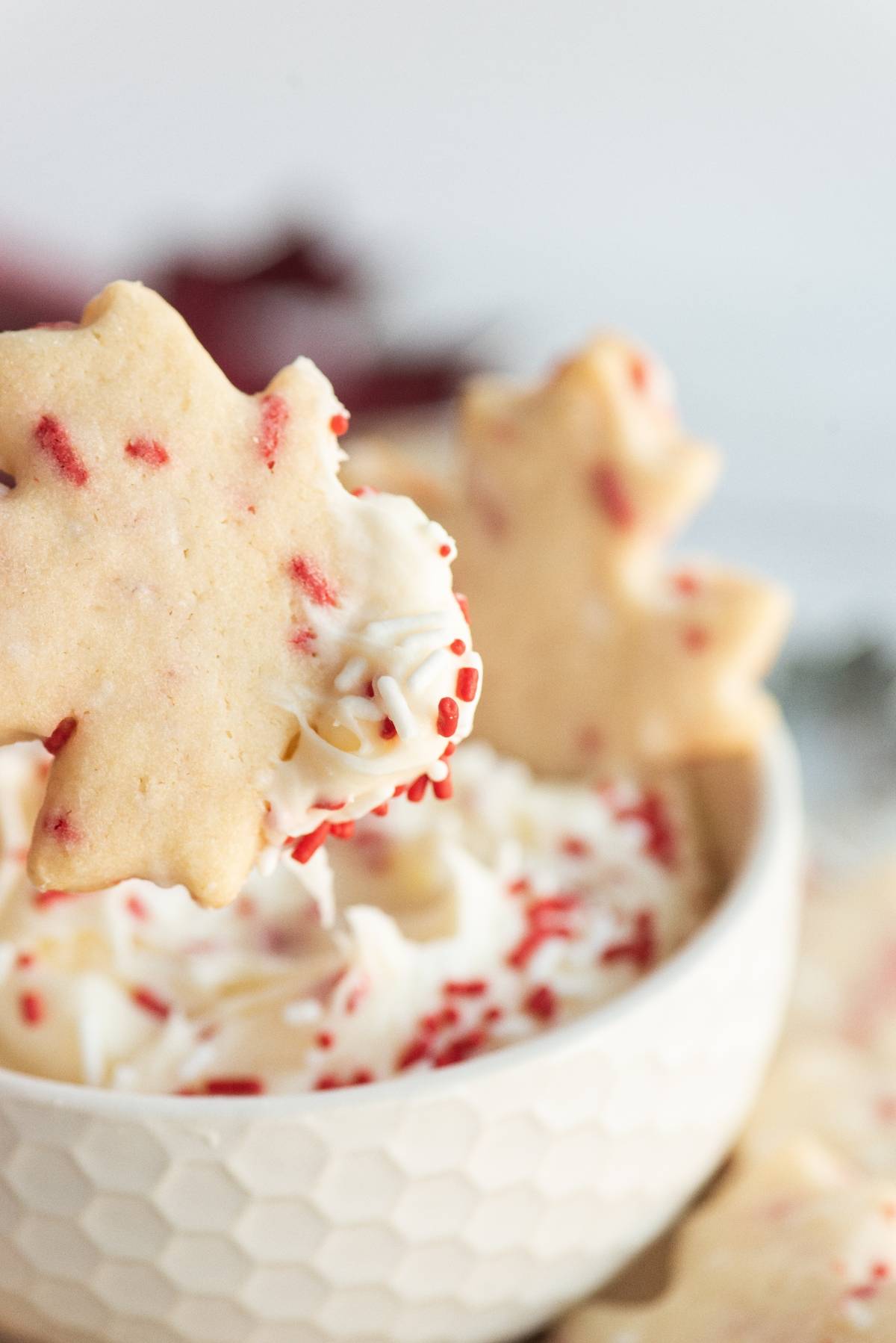 Maple leaf-shaped cookies with red sprinkles, dipped in a creamy white dip—perfect cookies to celebrate Canada Day or add to your list of recipes for a Canada Day party. Served in a textured white bowl for extra festive flair.