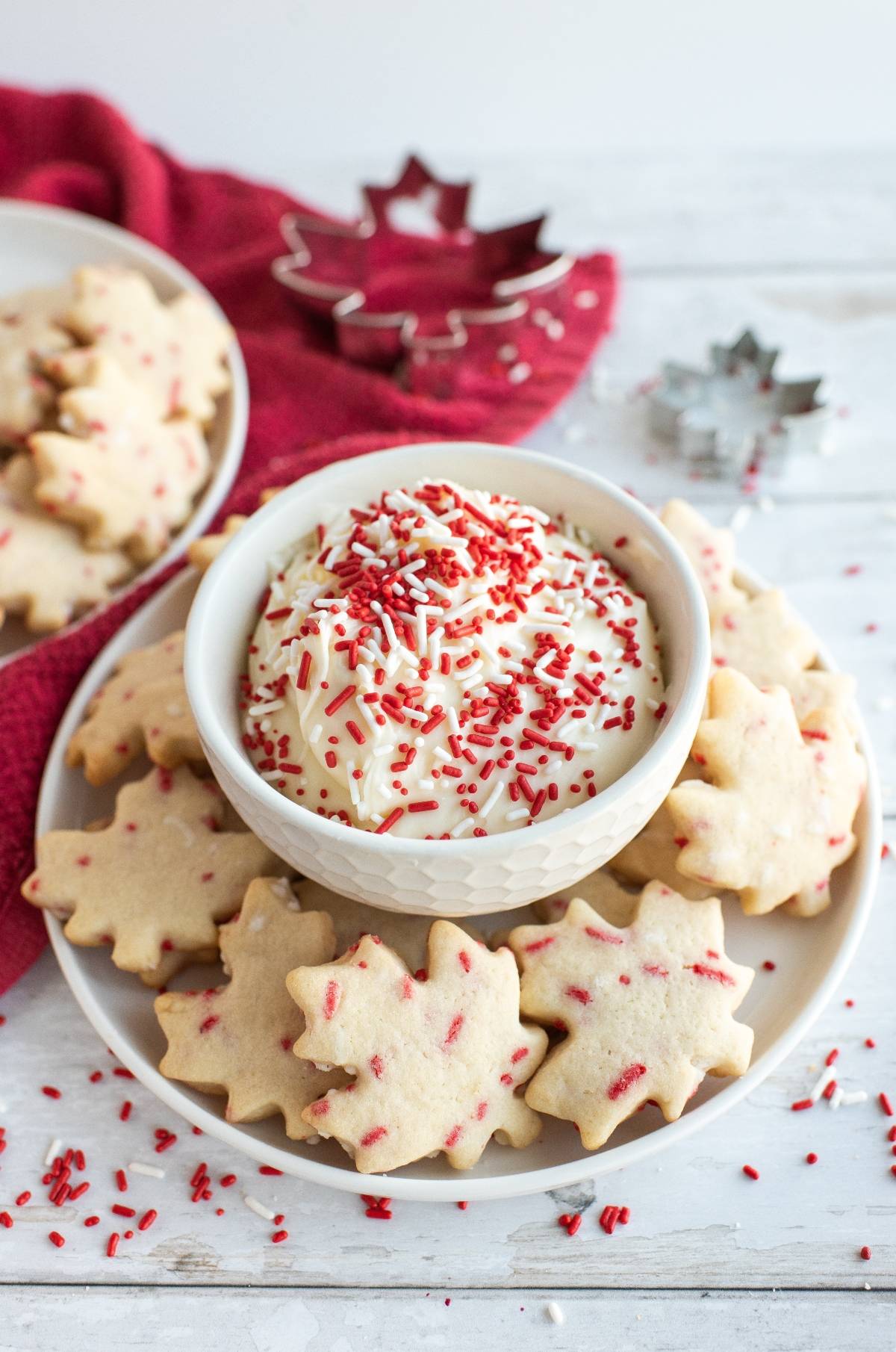 Plate of red and white Canada Day cookies with red sprinkles surrounds a bowl of white dip, perfect for Cookies to celebrate Canada Day.