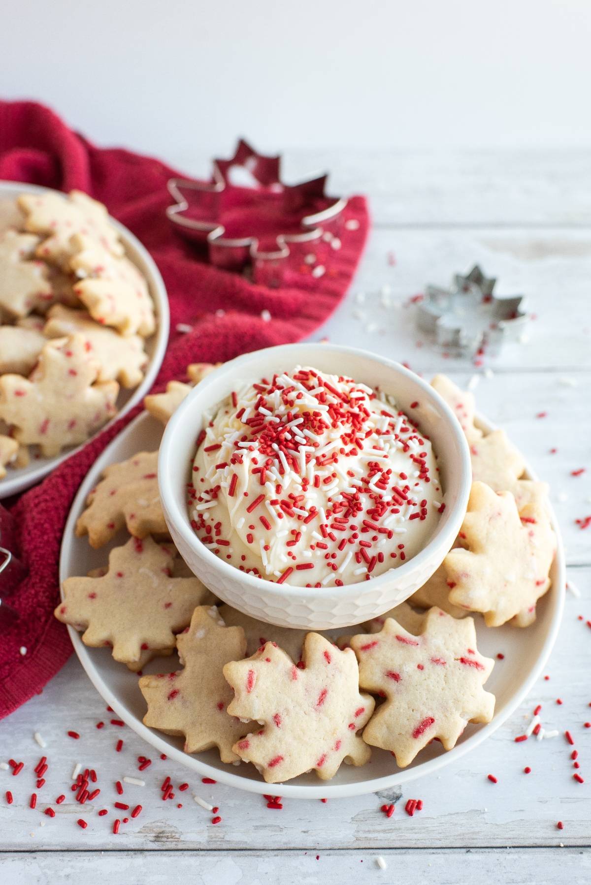Maple leaf cookies with red sprinkles surround a bowl of icing topped with red sprinkles on a white plate—perfect as festive Canada Day cookies for your celebration.