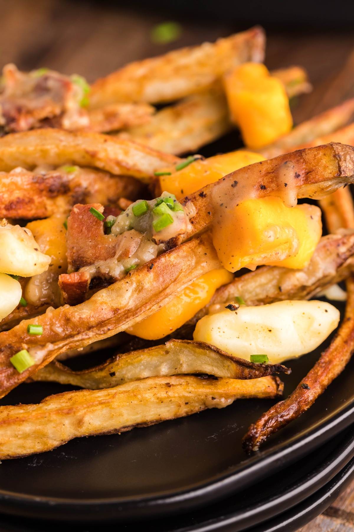 Close-up of loaded fries topped with melted cheese, bacon bits, and chopped green onions on a black plate—perfect for those craving a classic Canadian poutine or looking for inspiration for their next poutine recipe with meat.