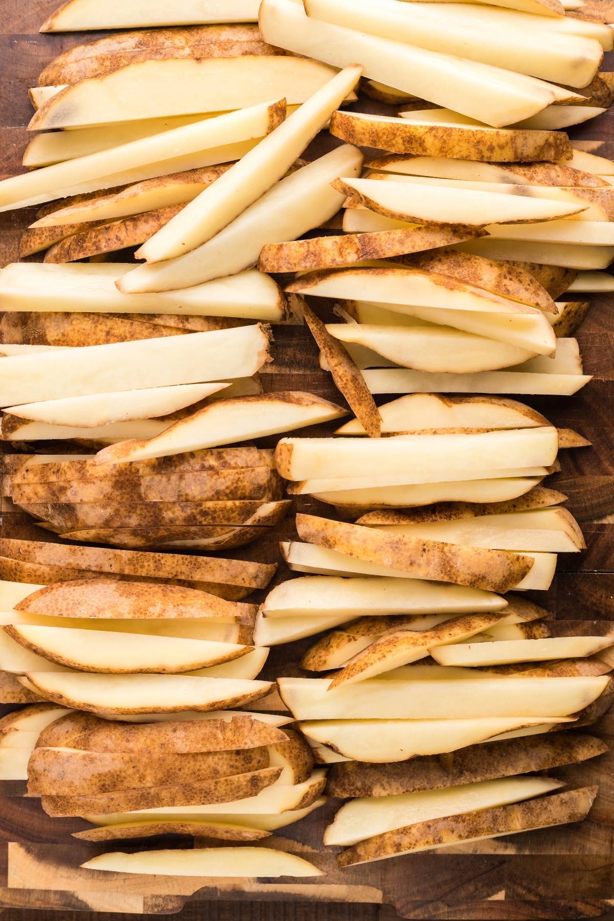 Raw russet potatoes sliced into thick strips on a wooden cutting board, perfect for making Canadian poutine or smothered fries with cheese curds and savory gravy.