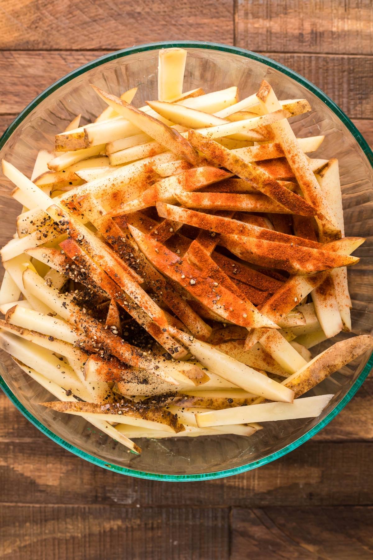 Raw potato strips in a glass bowl, sprinkled with spices and black pepper, perfect for starting your homemade Canadian poutine or exploring how to make poutine at home.
