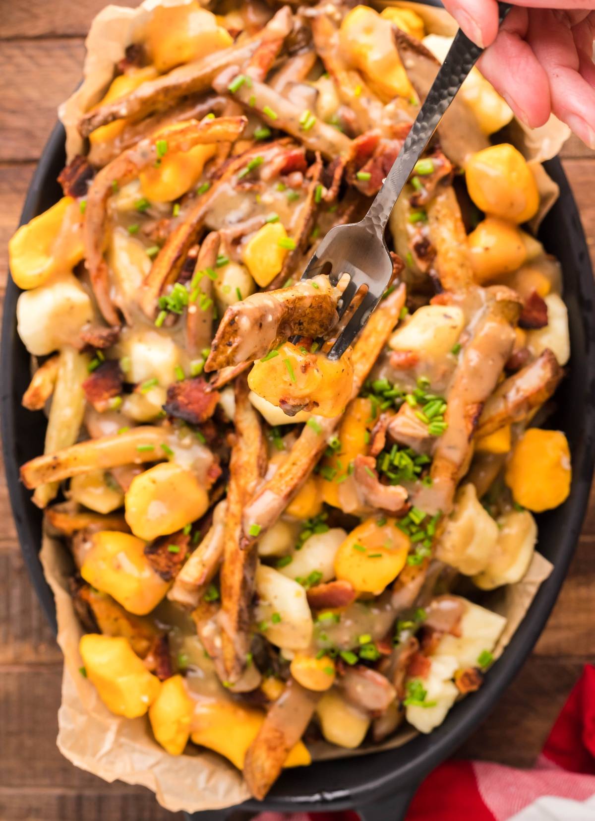 A hand holds a fork over a plate of Canadian poutine with fries, cheese curds, gravy, bacon, and chives—capturing the essence of how to make poutine at home.