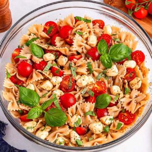 Glass bowl of Caprese pasta salad with cherry tomatoes, mozzarella, basil, and herbs on a white surface.