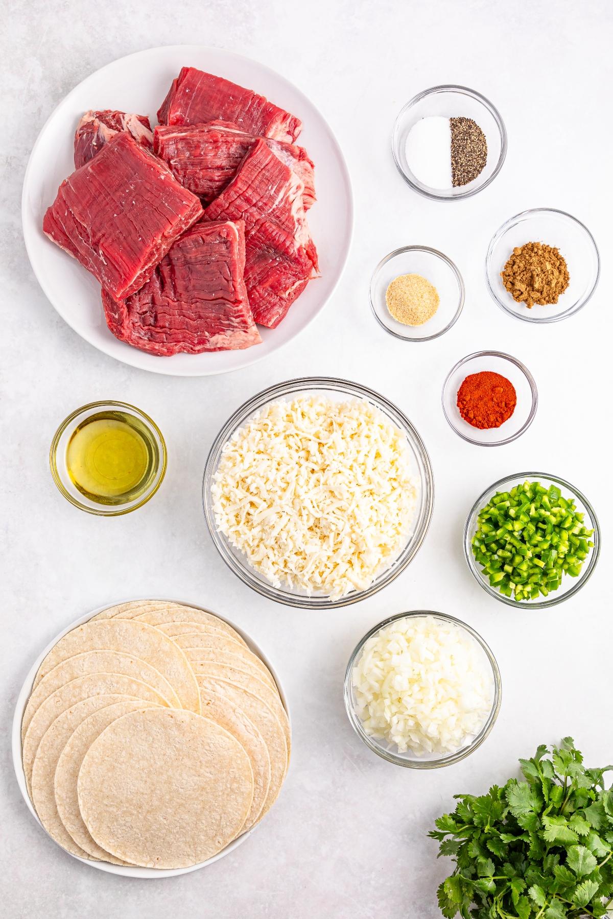 Raw beef, shredded cheese, tortillas, spices, oil, chopped onion, peppers, and fresh cilantro on a white surface.