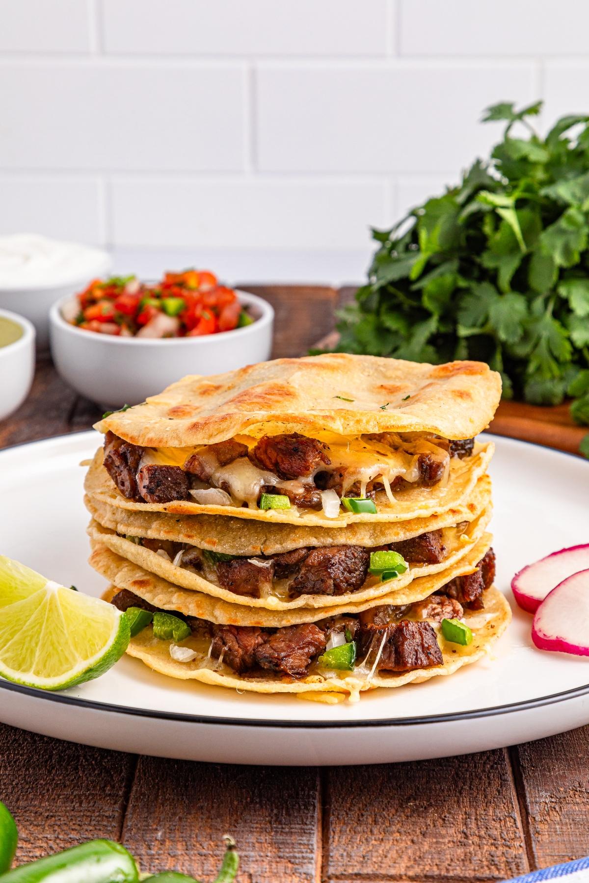 Three quesadillas stacked on a plate with beef, cheese, and green onions, garnished with lime and radish slices.